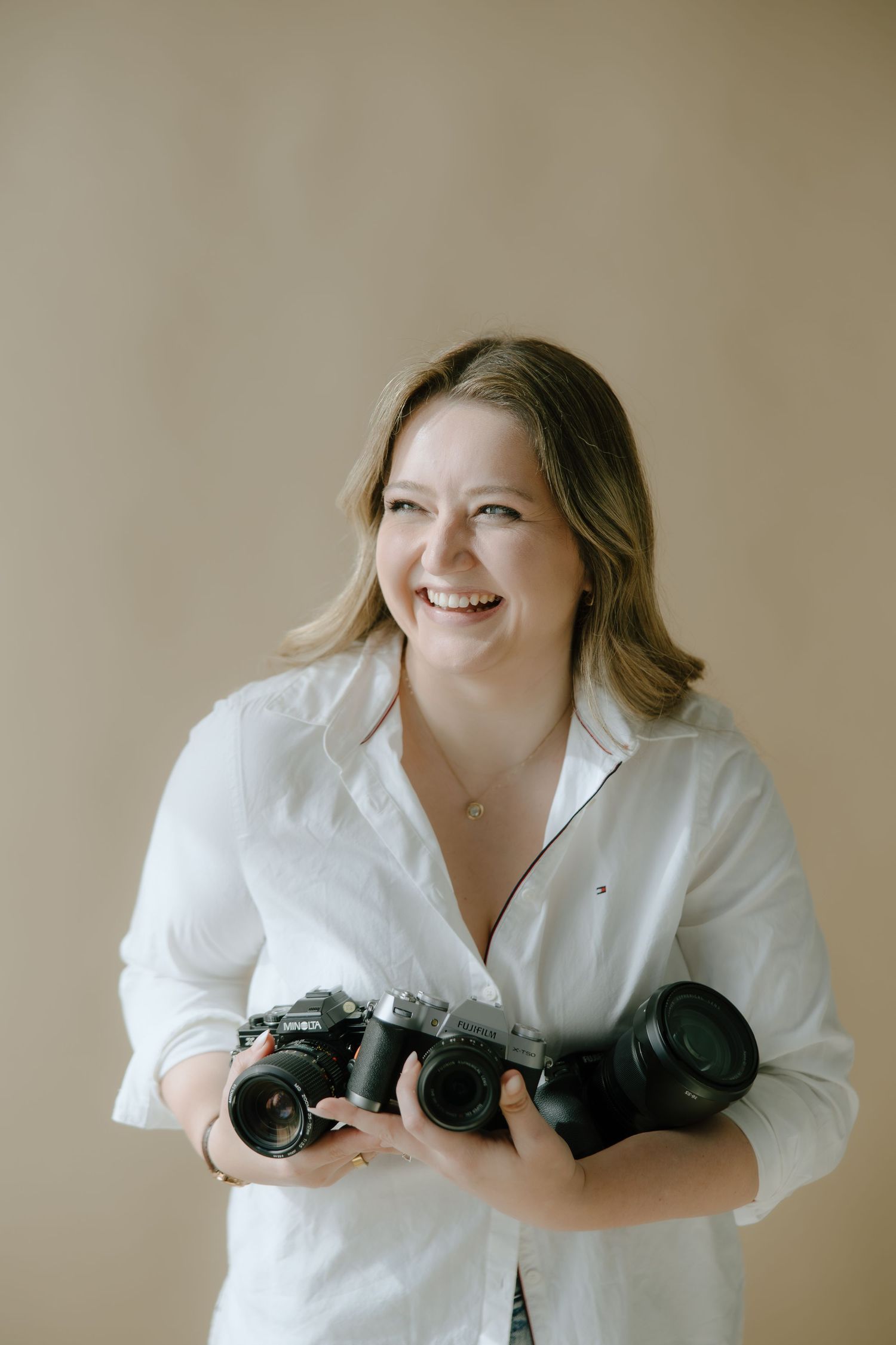 Smiling woman in white shirt holding multiple professional cameras against a neutral beige background.