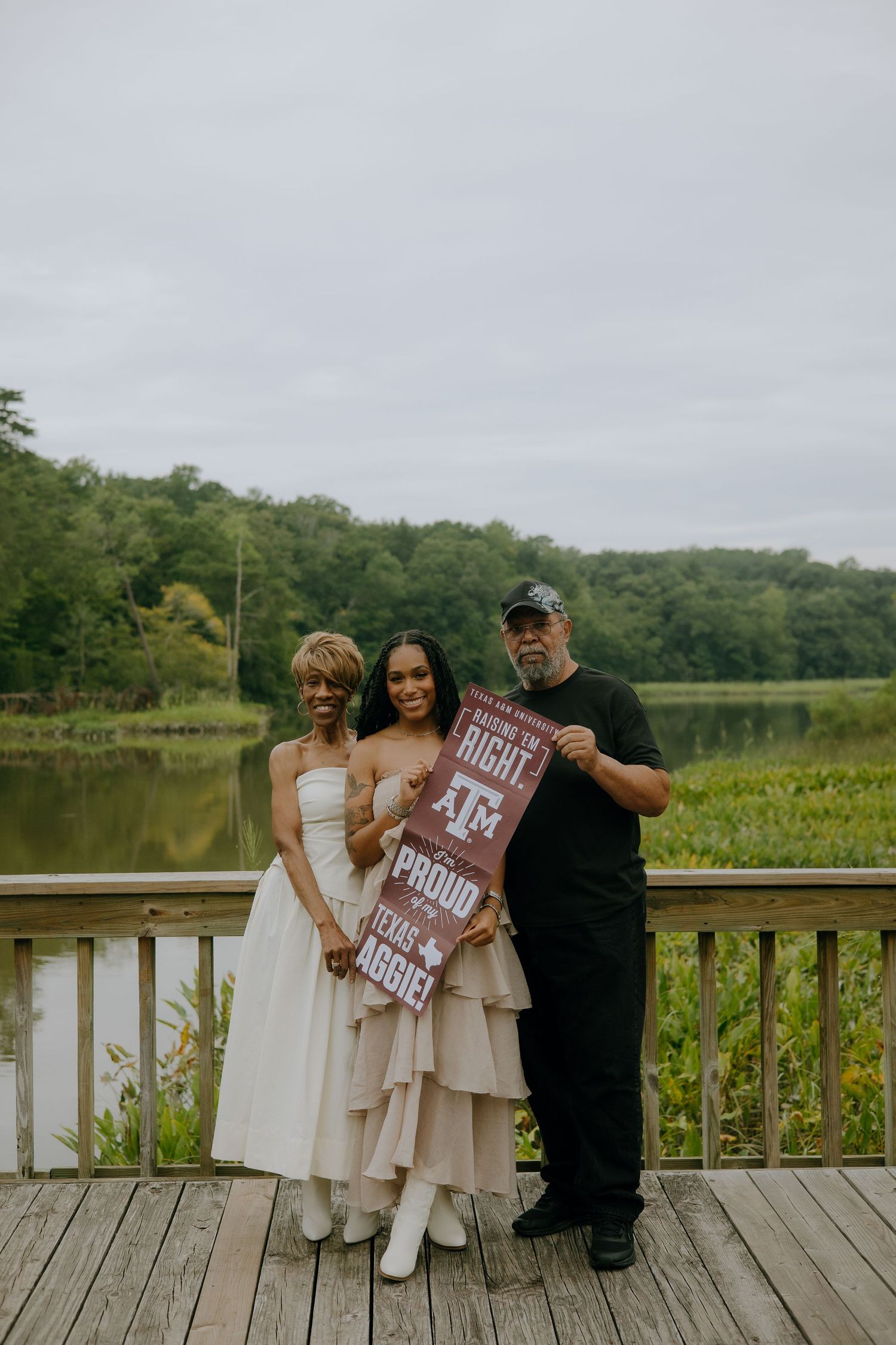 Family of three posing on wooden deck holding Texas A&M Aggies sign near scenic lake and green trees.