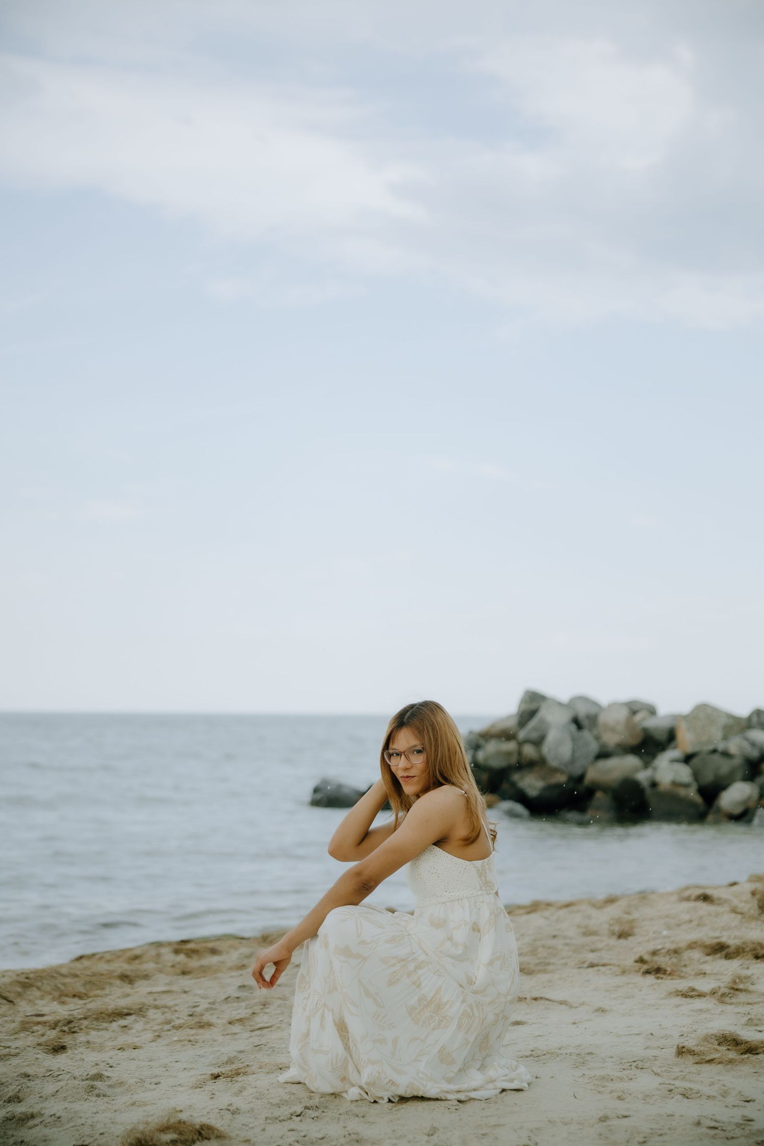 Young woman in white floral dress sitting on sandy beach with rocky shoreline and calm ocean in background.