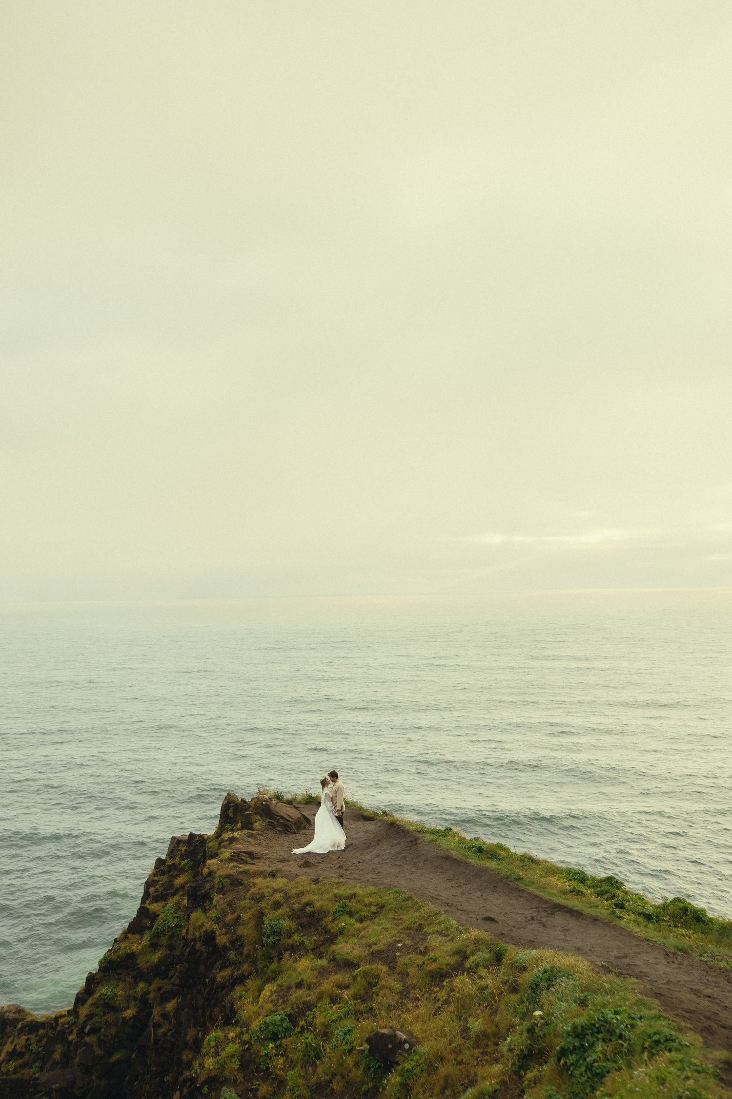 A couple in wedding attire stands on a dramatic coastal cliff overlooking a calm ocean under a soft, overcast sky.