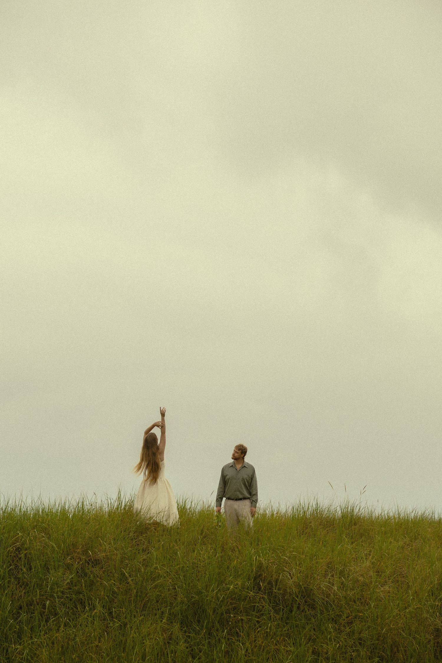 Two people standing on a grassy hill under a cloudy sky, one with long hair raising their arm.