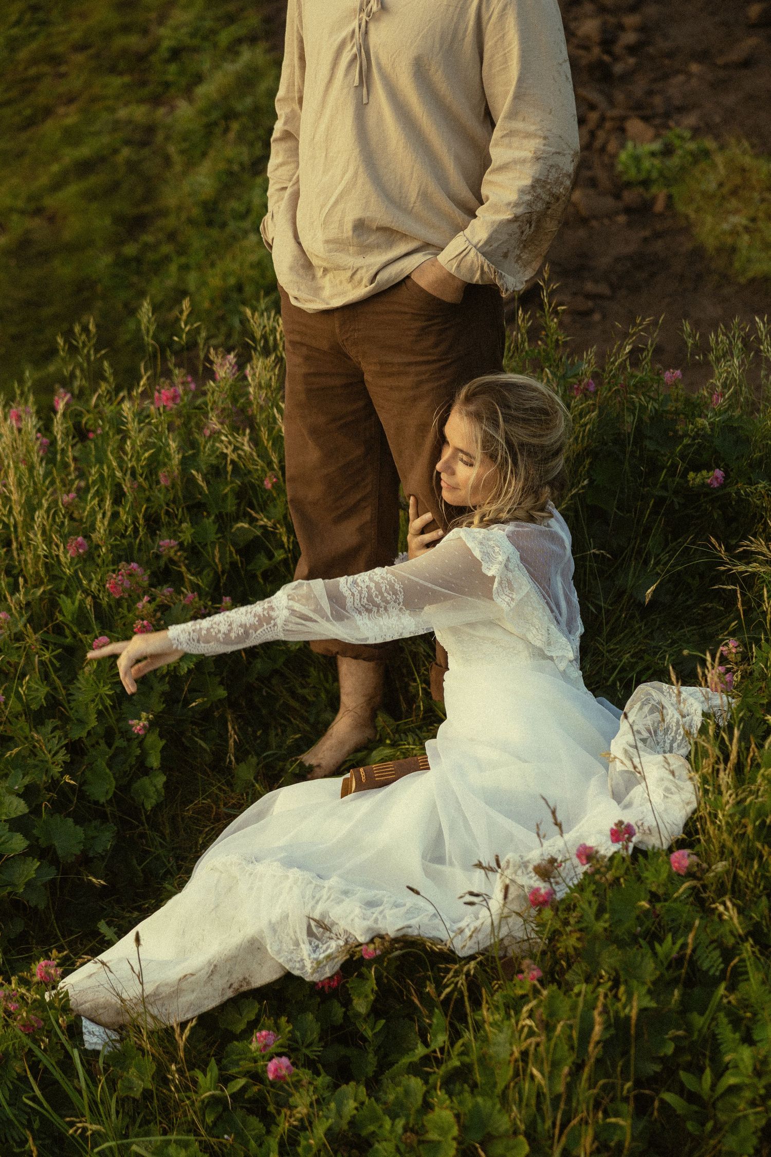 Bride in a white lace gown sitting among wildflowers beside her groom in a sunlit meadow.