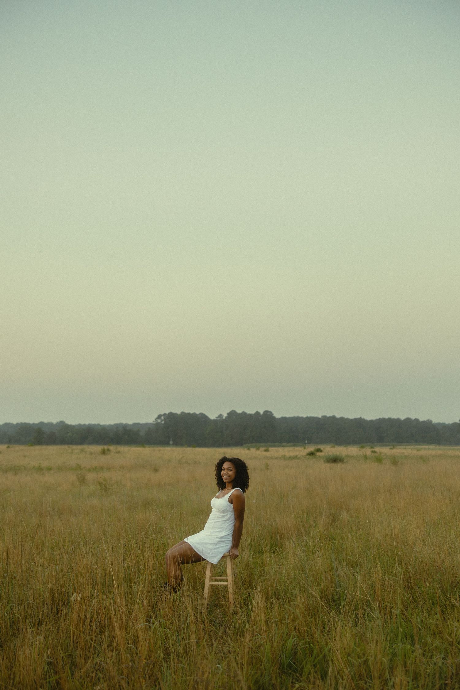 A young Black woman in a white dress sits on a stool in a golden field at dusk.