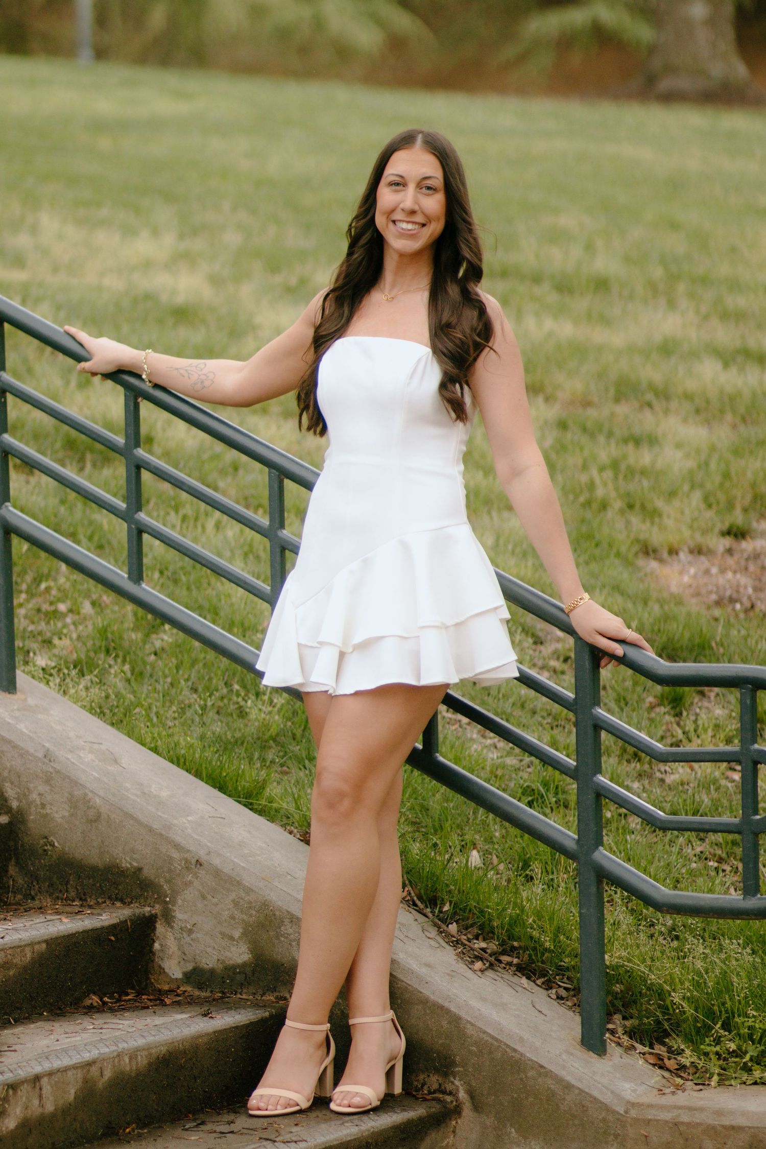 Graduate in white dress poses on concrete steps with metal railing outdoors.