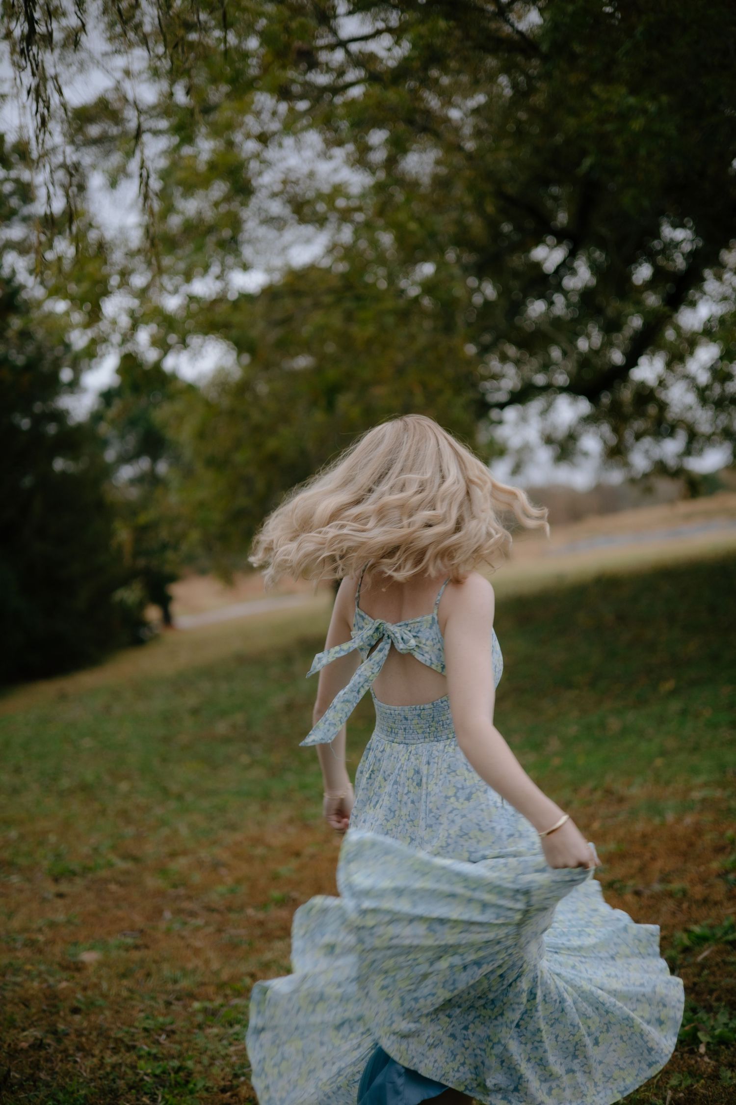 A woman in a flowy blue floral dress twirls outdoors on a grassy hill with autumn trees in the background.