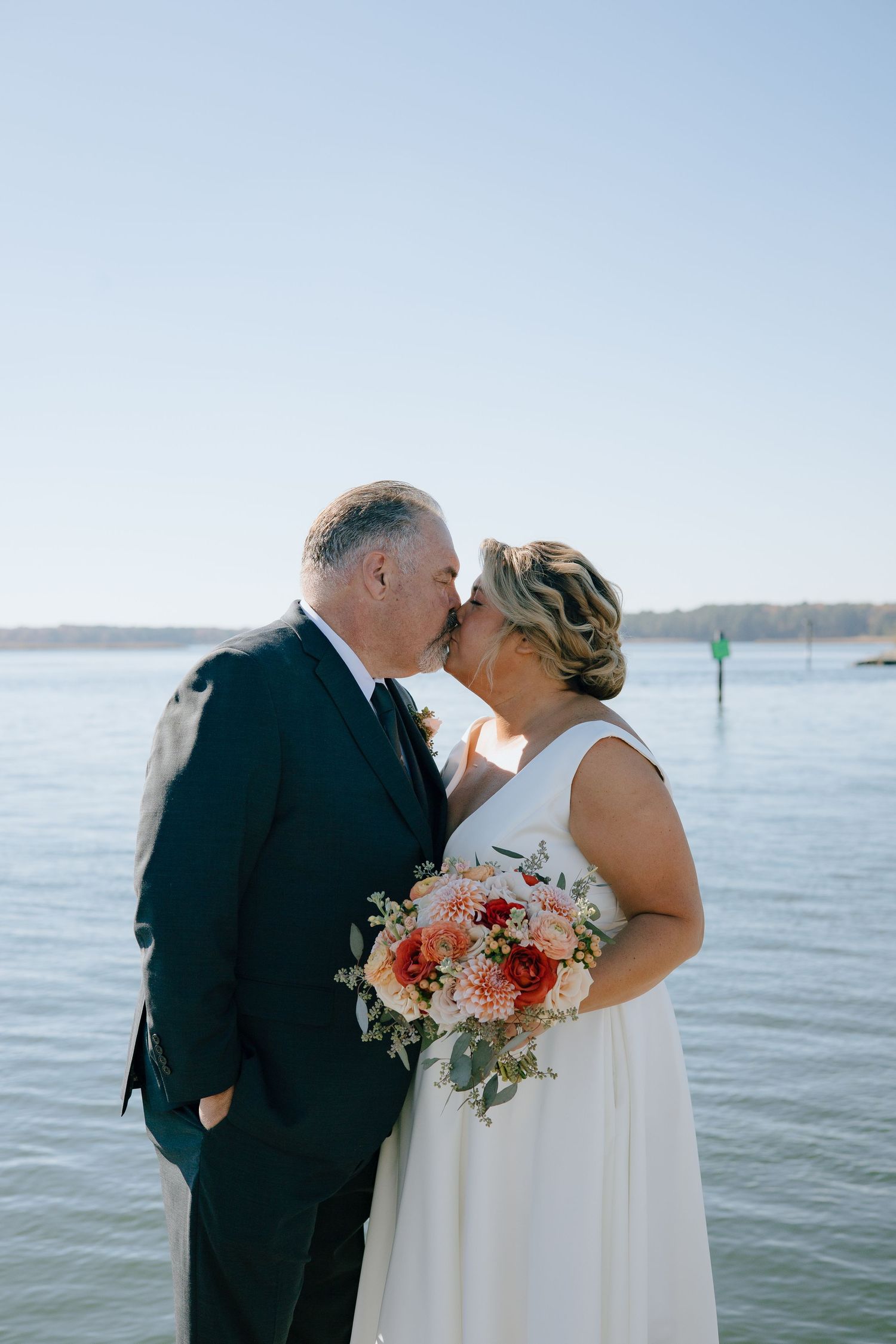 A tender moment between newlyweds by the waterfront with a beautiful coral bouquet.