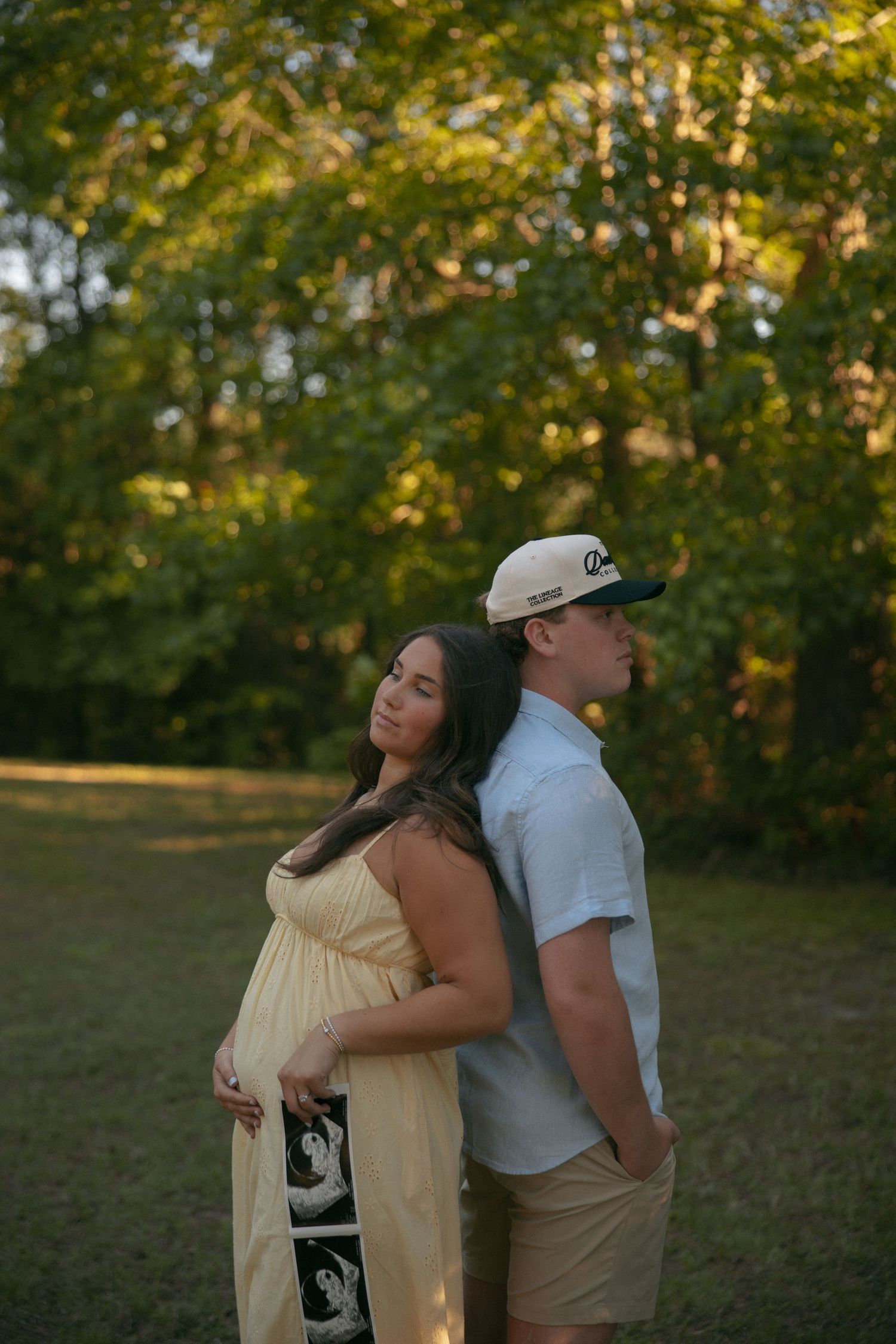 Pregnant woman in yellow dress holds ultrasound photos while standing back-to-back with partner outdoors at golden hour.