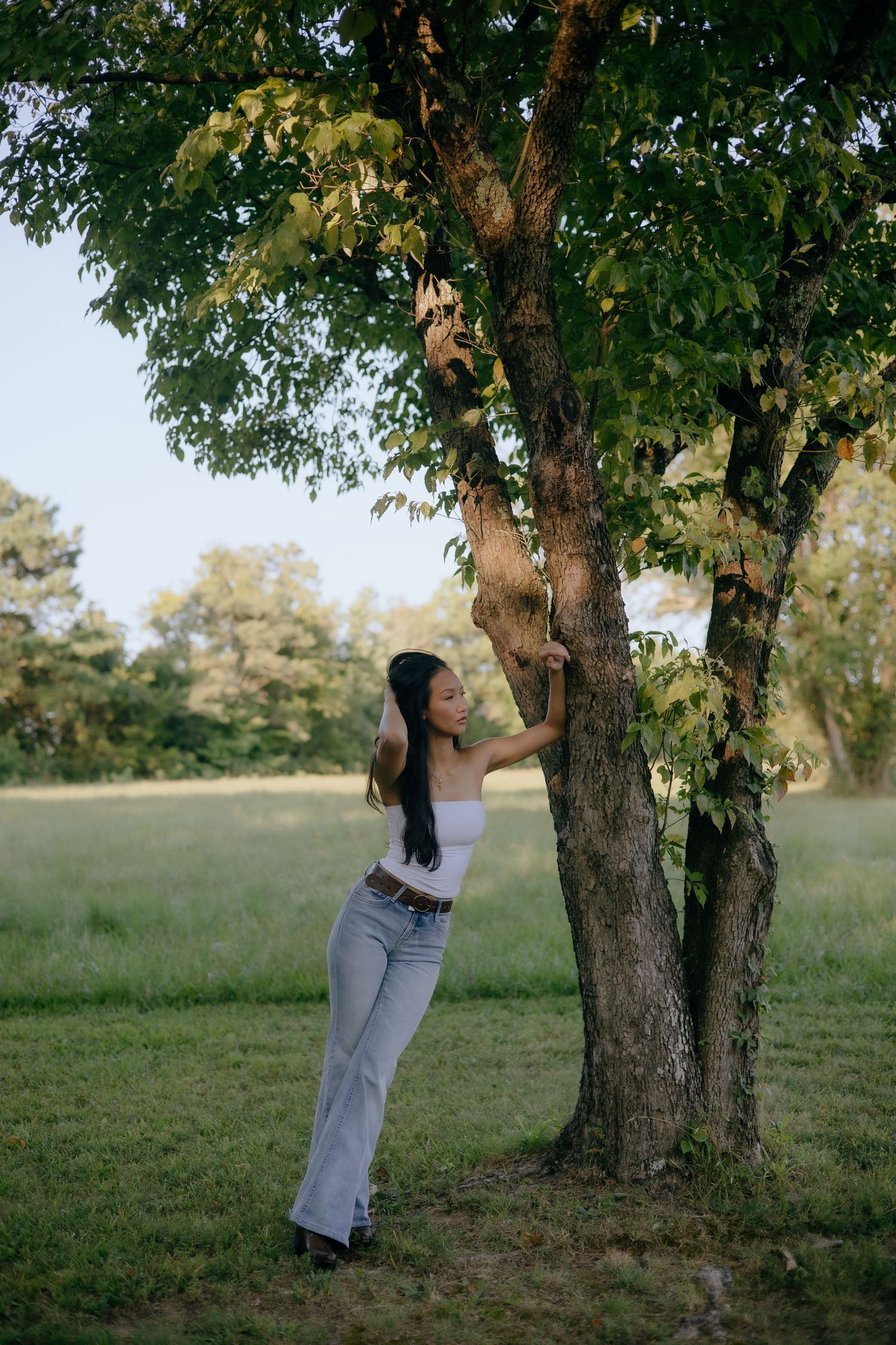 Young woman in white top and jeans leaning against a tree in a lush green park during golden hour.