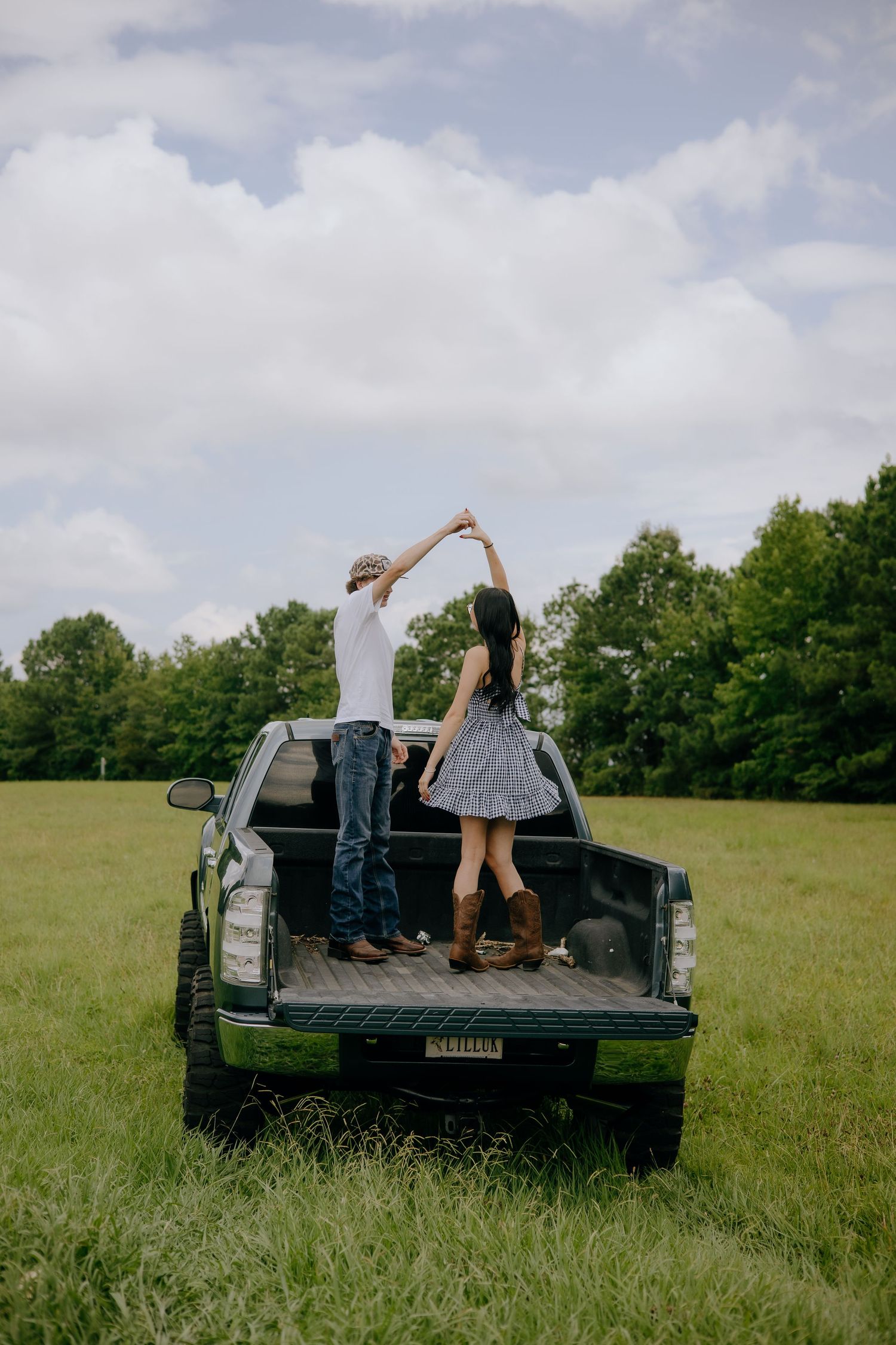 Couple dancing in the bed of a green pickup truck in a lush green field under a cloudy sky.