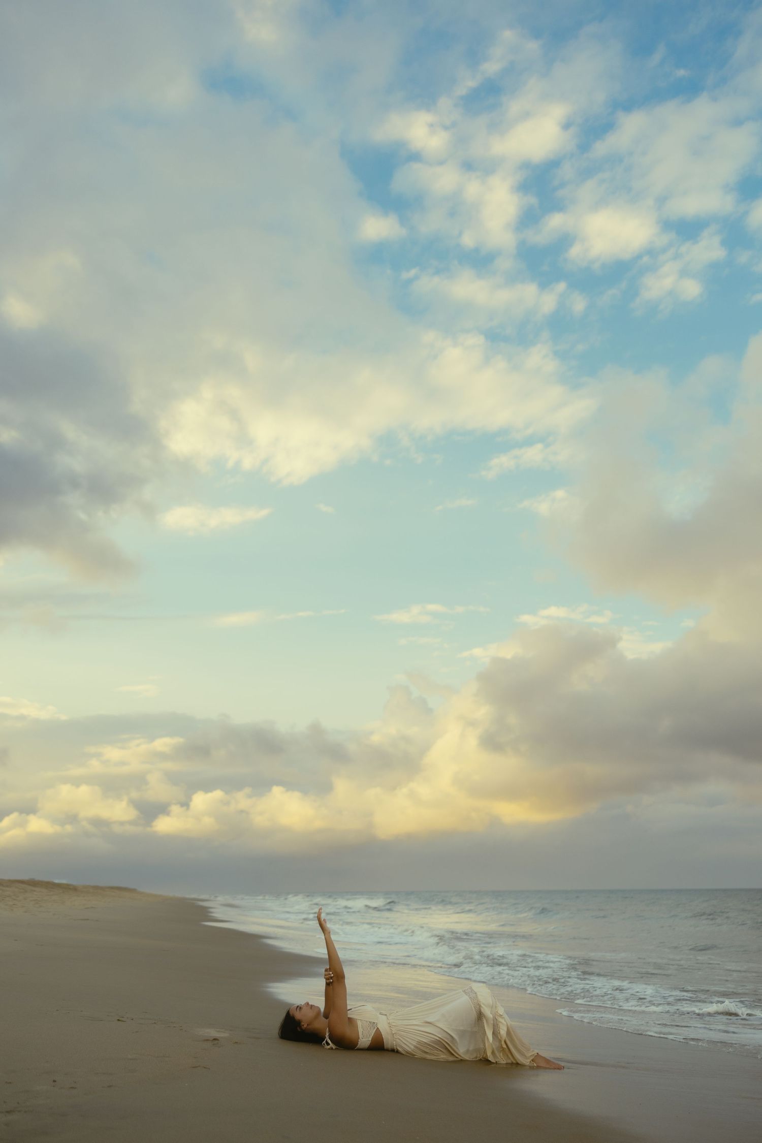 Woman in white dress lying on sandy beach with arm raised, dramatic cloudy sky at sunset over ocean waves.