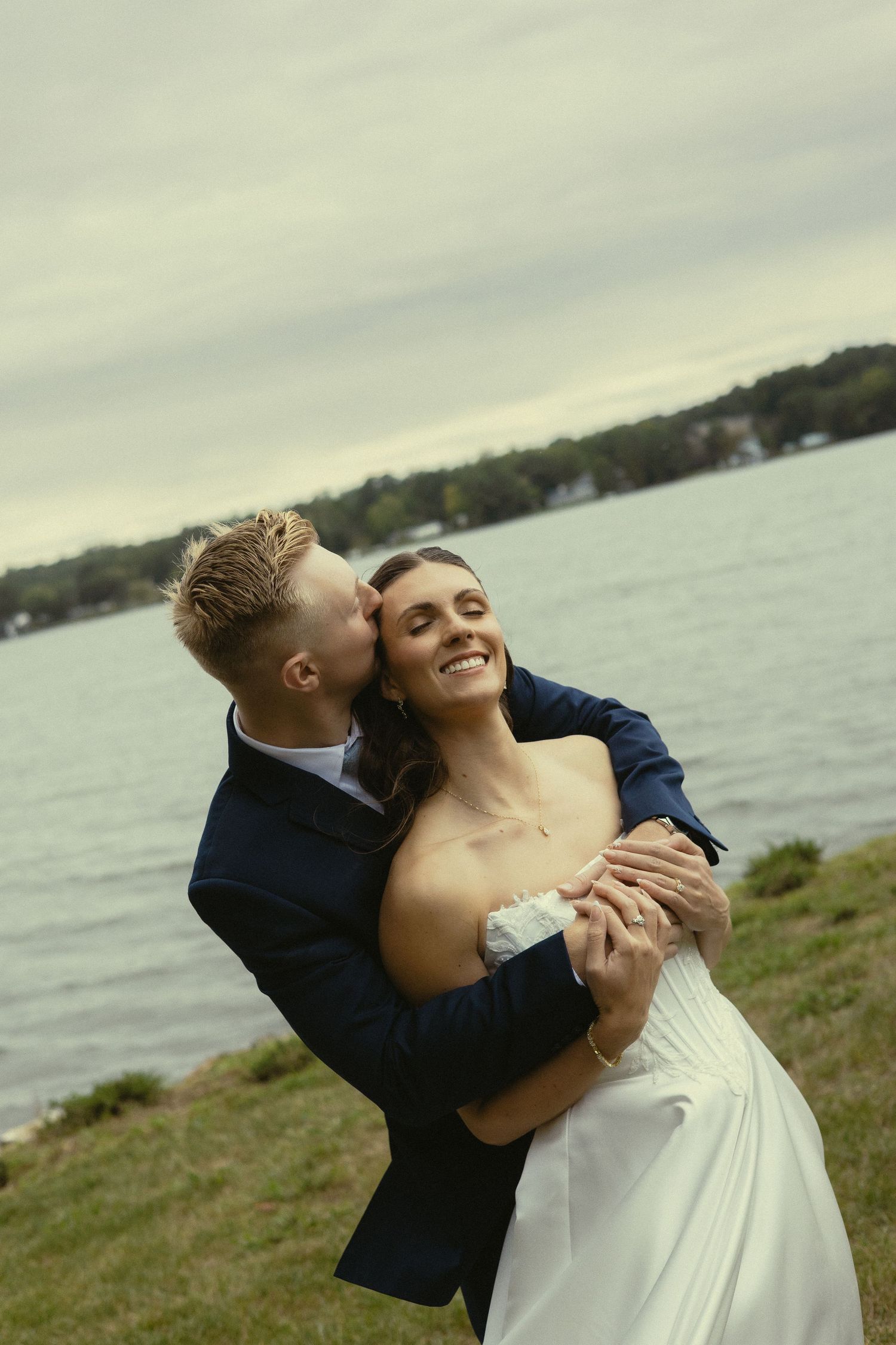 Groom in navy suit kissing bride on cheek while embracing her outdoors near a lake on their wedding day.
