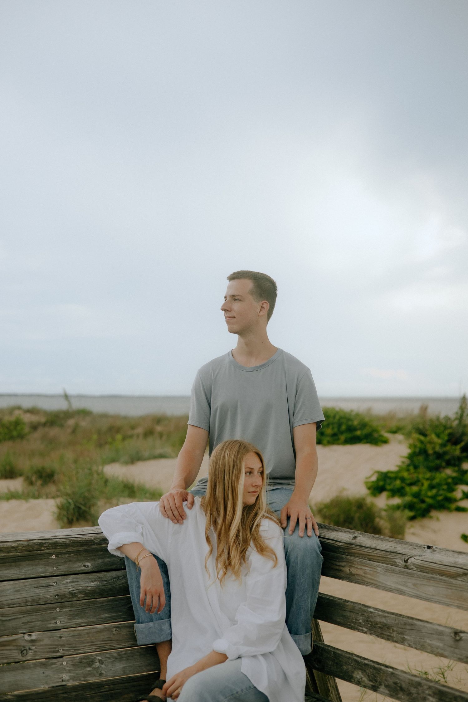 A couple sits together on a wooden deck overlooking a coastal landscape on a cloudy day.