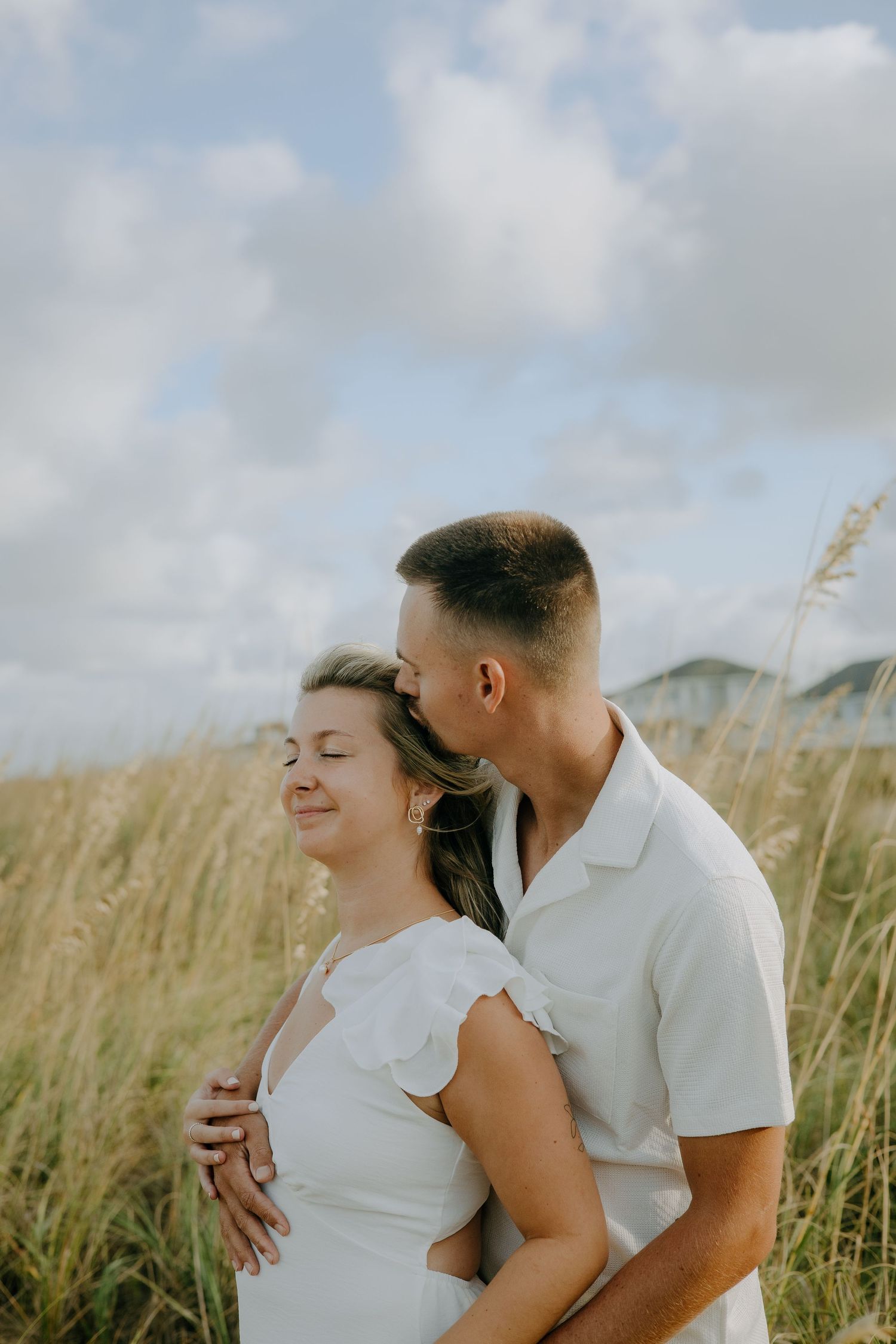 A tender moment shared between a couple in white clothing among tall beach grass.