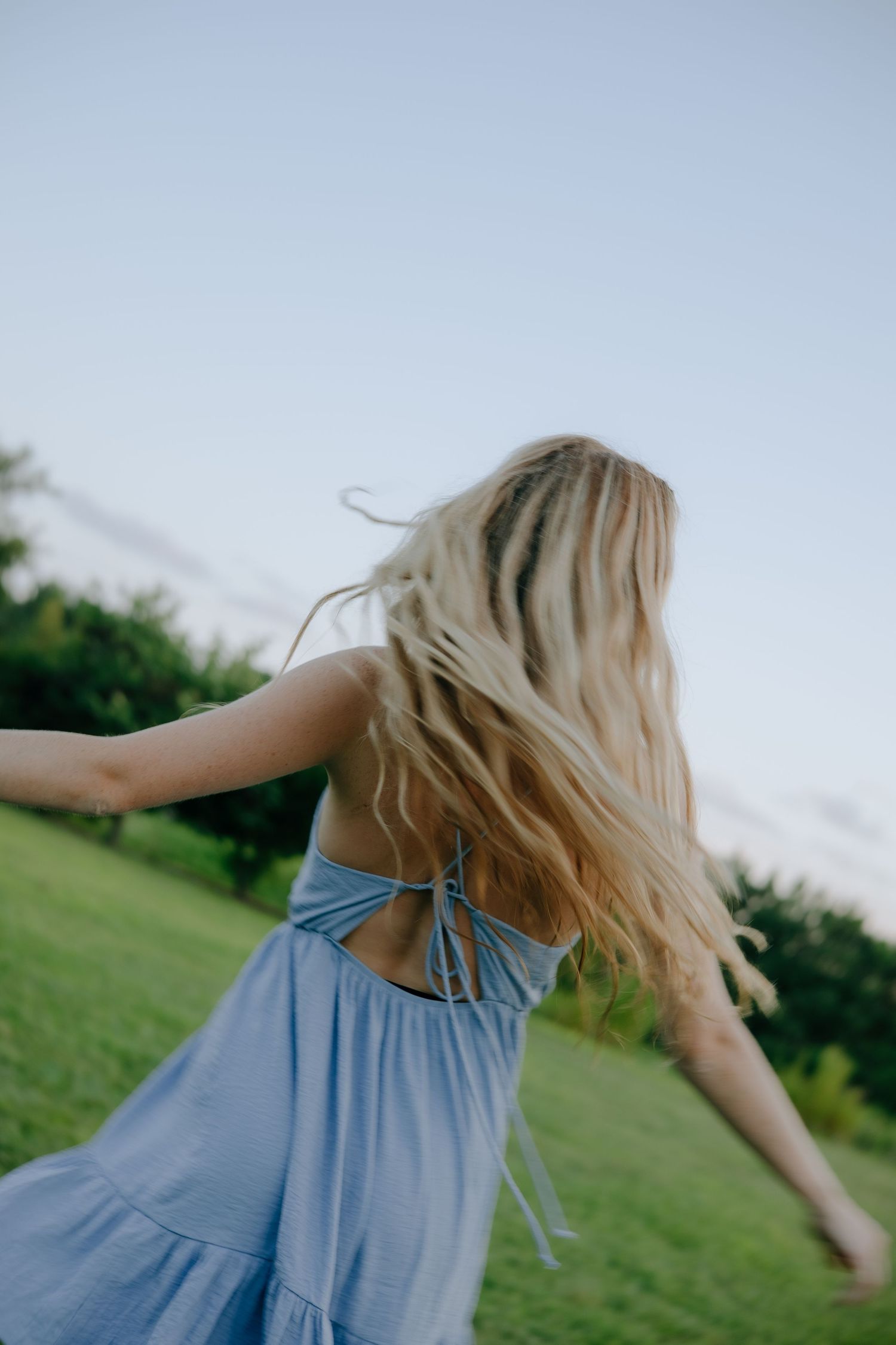 A person in a light blue sundress spins joyfully in a grassy field with long blonde hair flowing in the breeze.