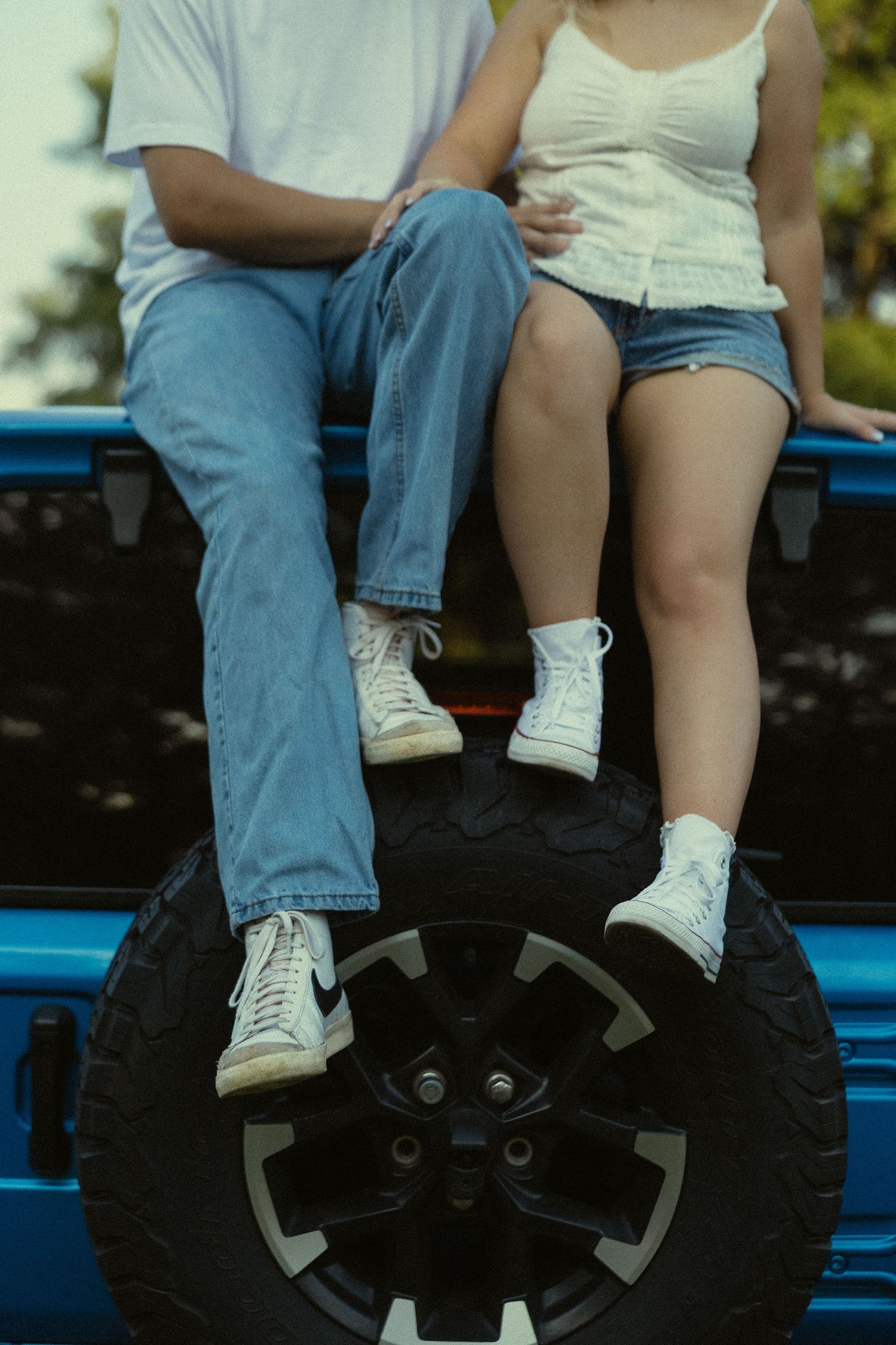 Couple sitting on blue Jeep spare tire wearing jeans and white sneakers on a sunny day.
