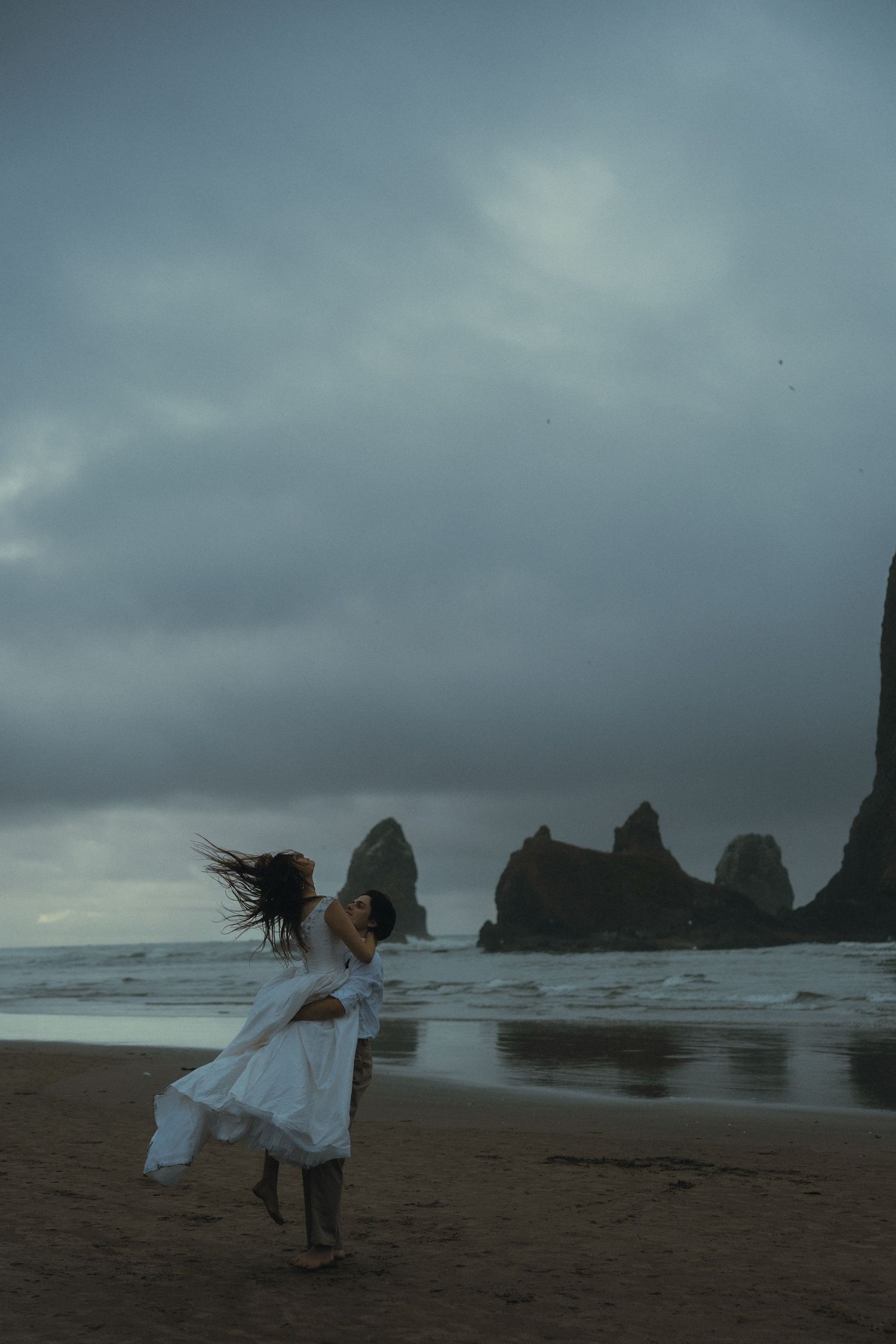 Two people in white dresses embrace on a moody beach with dramatic sea stacks and stormy skies.