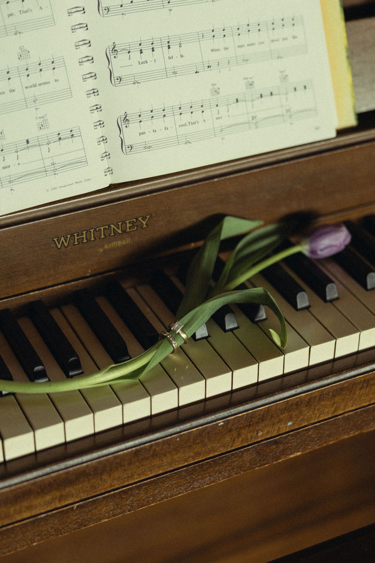 A purple tulip and ring rest on Whitney piano keys beside open sheet music.