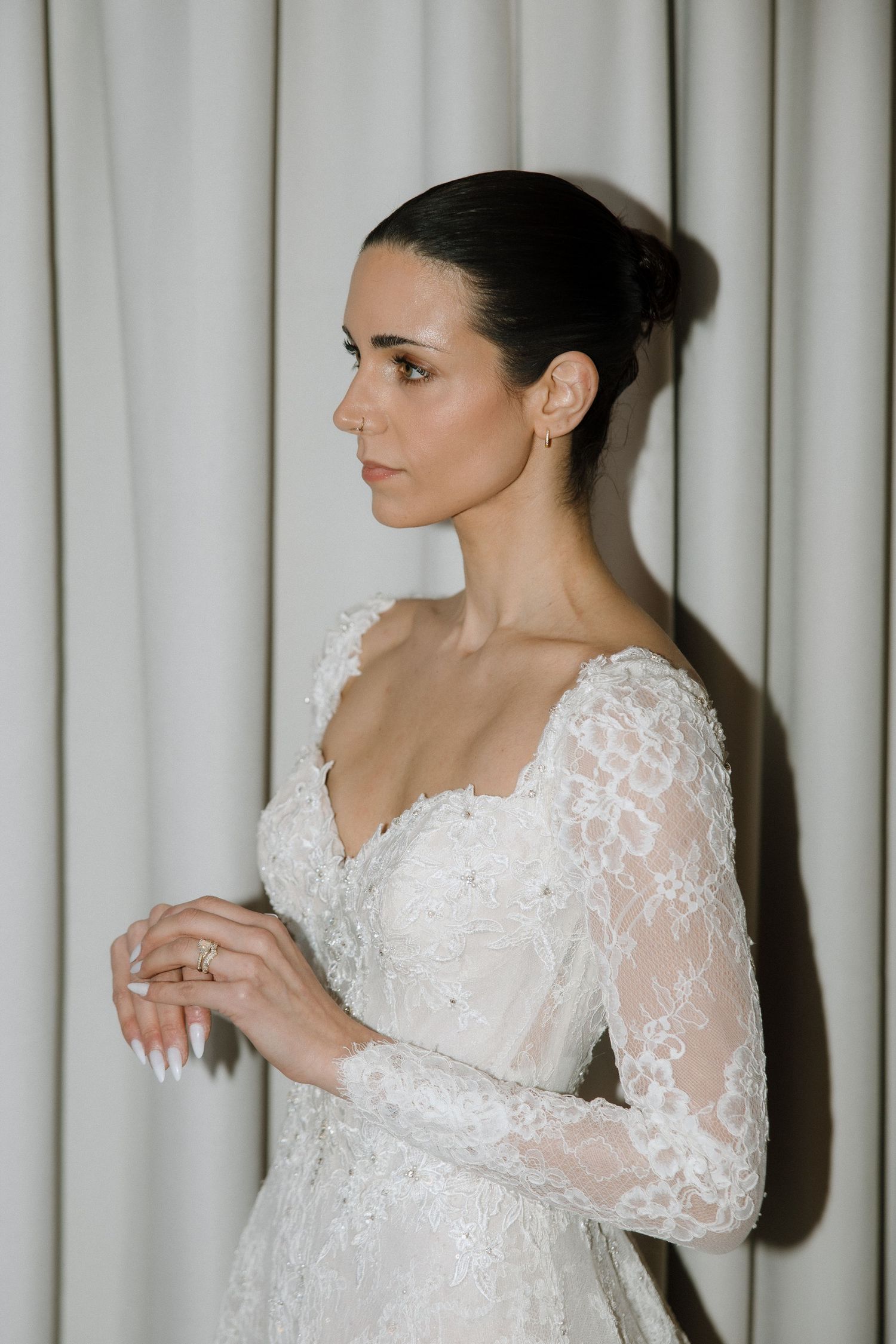 Bride in elegant long-sleeve lace wedding gown with V-neckline, hair in updo, posing against white curtains.