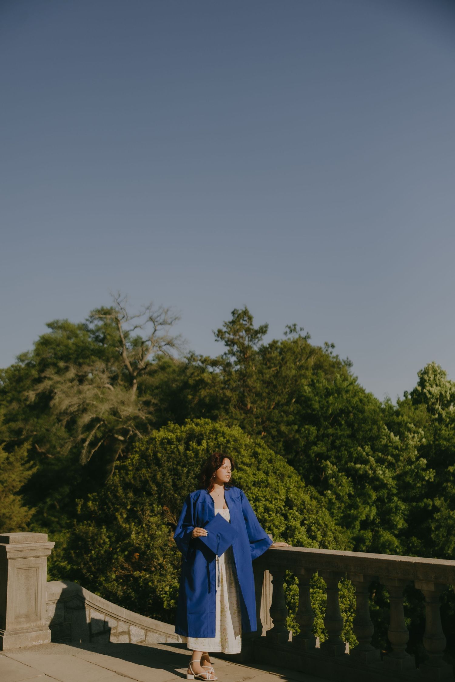 Woman in blue coat and white dress stands on stone balcony surrounded by lush green trees under clear blue sky.