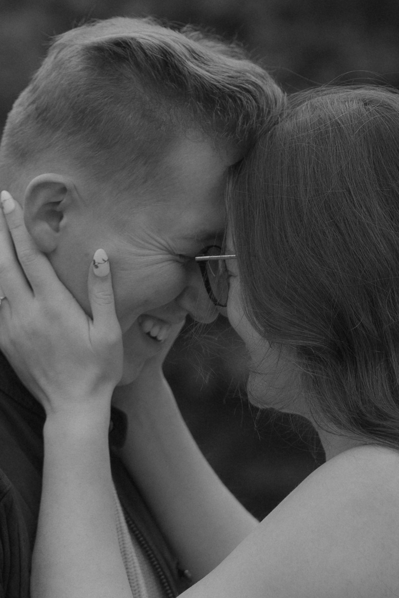 Black and white photo of a smiling couple touching foreheads, woman with glasses holding mans face tenderly.