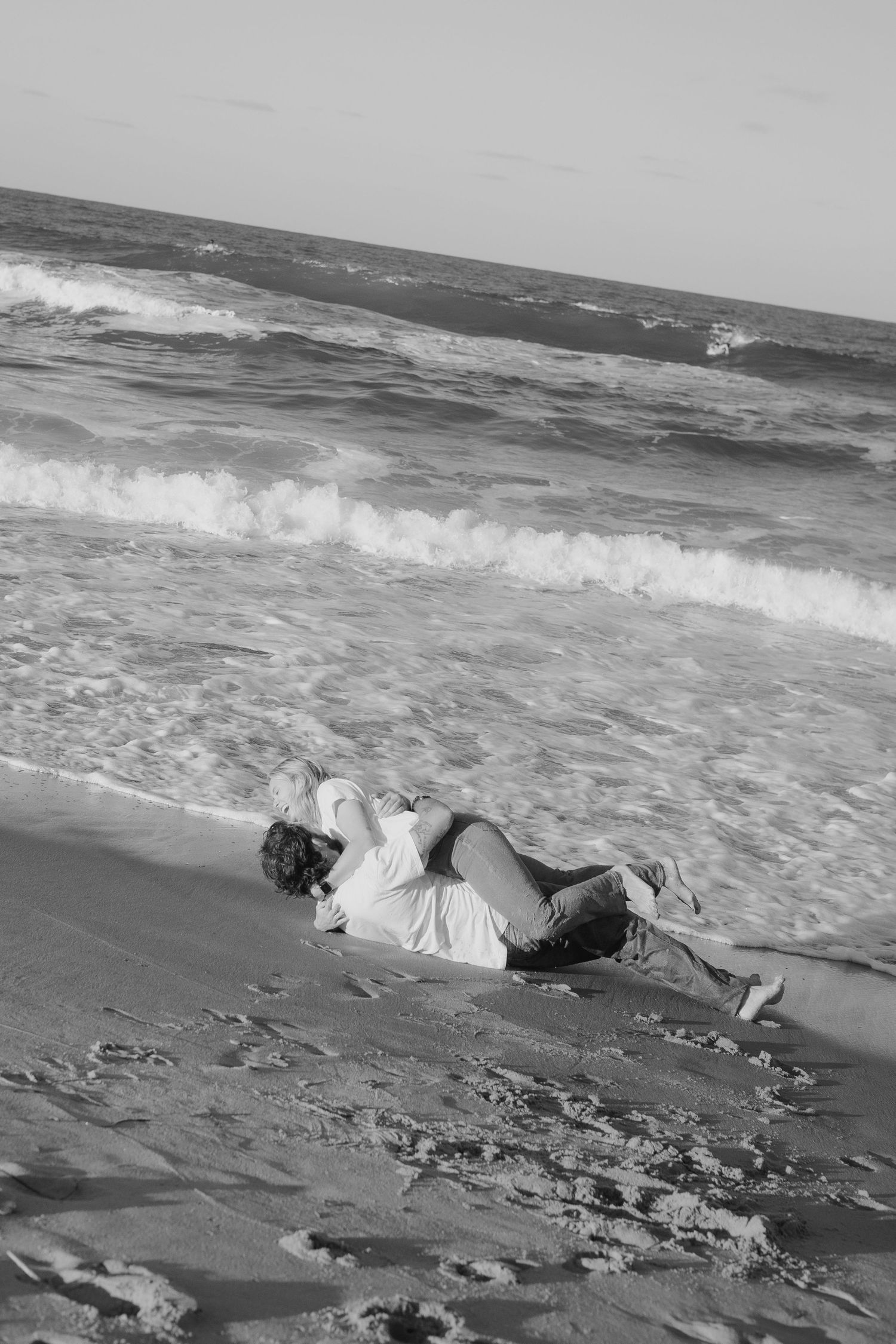 A couple embraces on a sandy beach as ocean waves crash onto the shore in black and white.