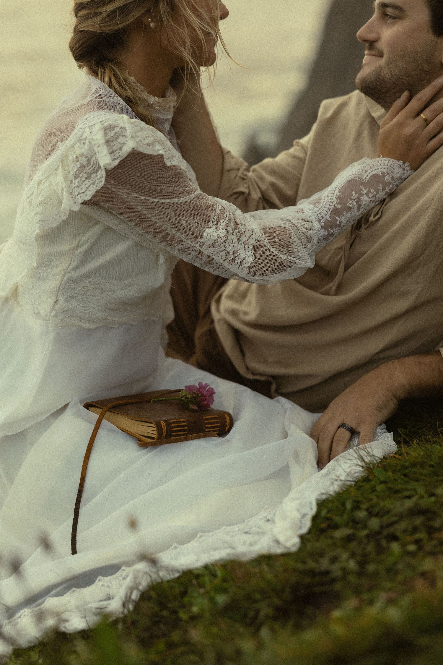 Bride in lace wedding dress sits with groom on mossy ground, gently touching his face in a romantic outdoor setting.
