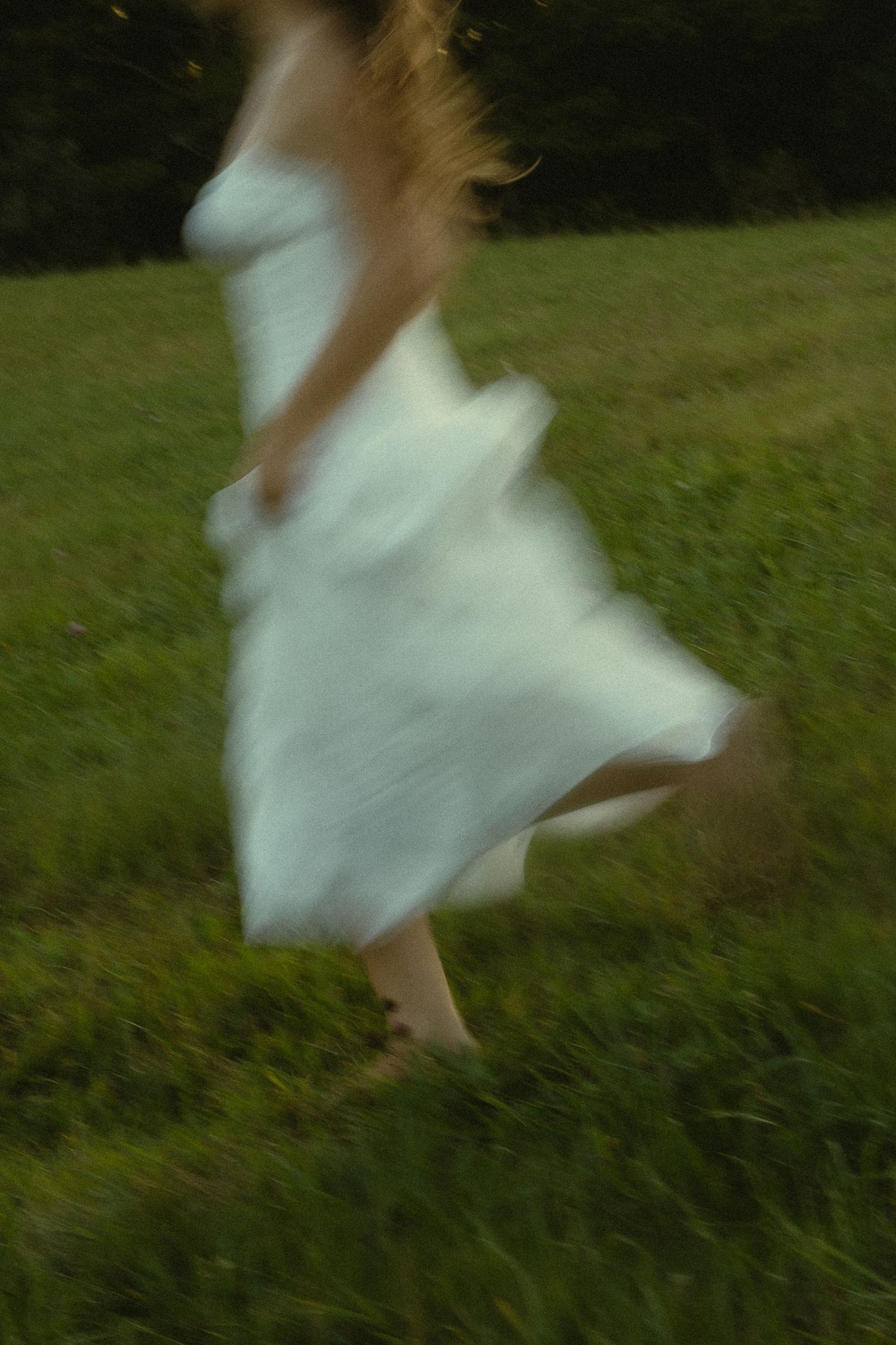 A blurred woman in a white sundress runs barefoot through a lush green meadow, captured in motion.