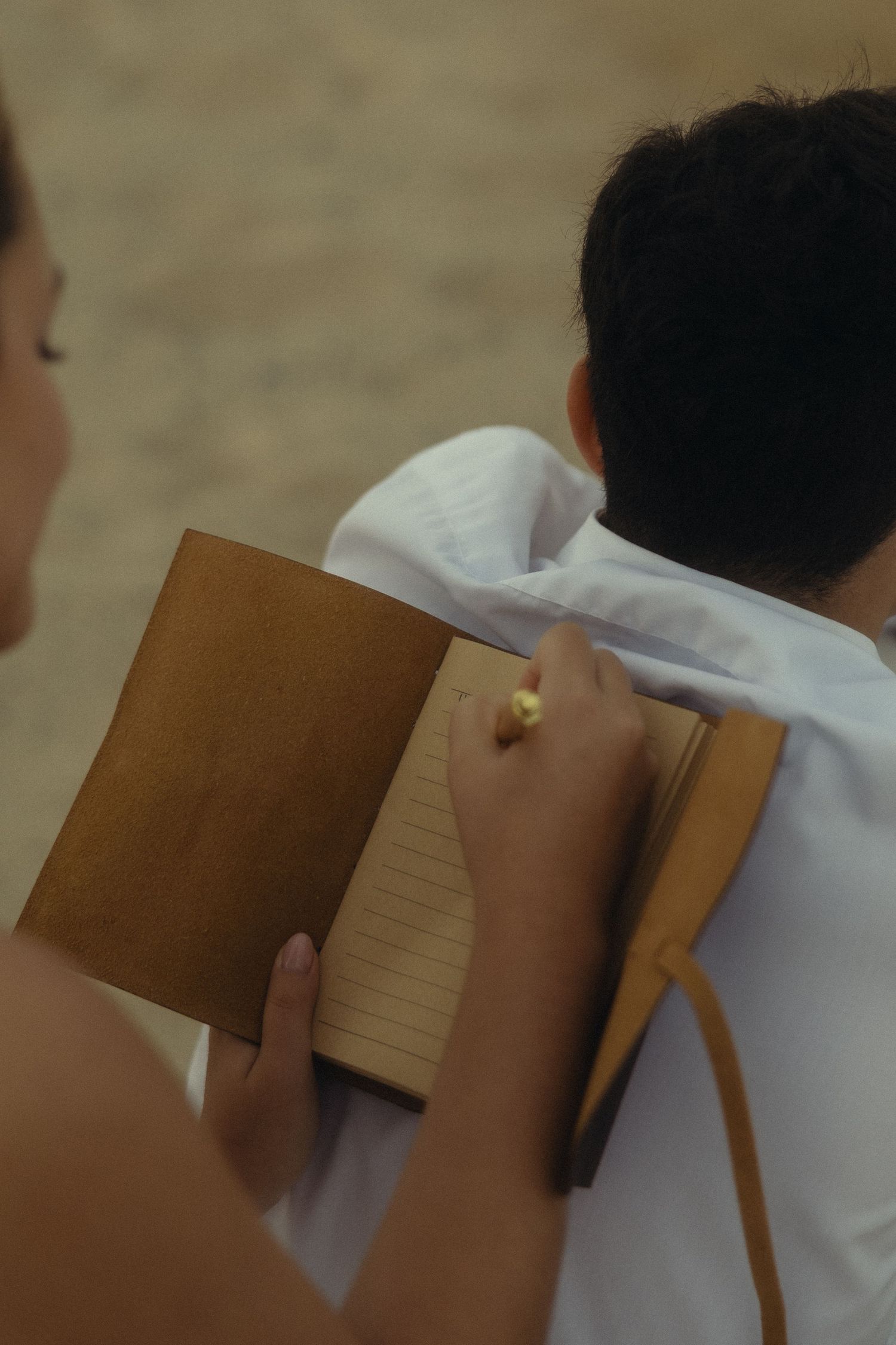 Person writing in a brown leather journal with a gold pen, viewed from behind another person in white shirt.