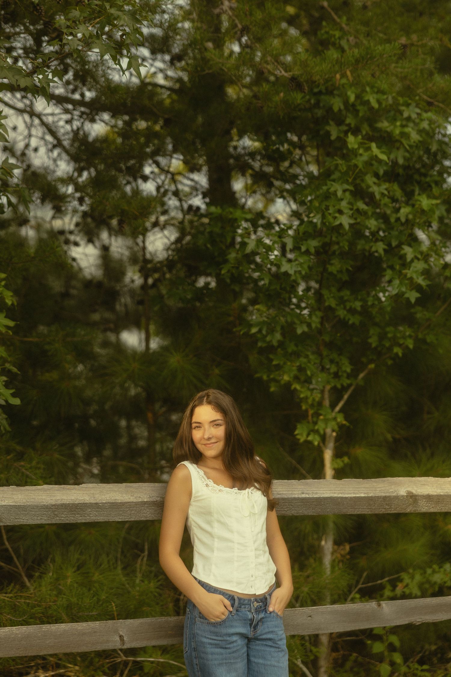 Young woman in white tank top and jeans smiling by a wooden fence with lush green trees in background.