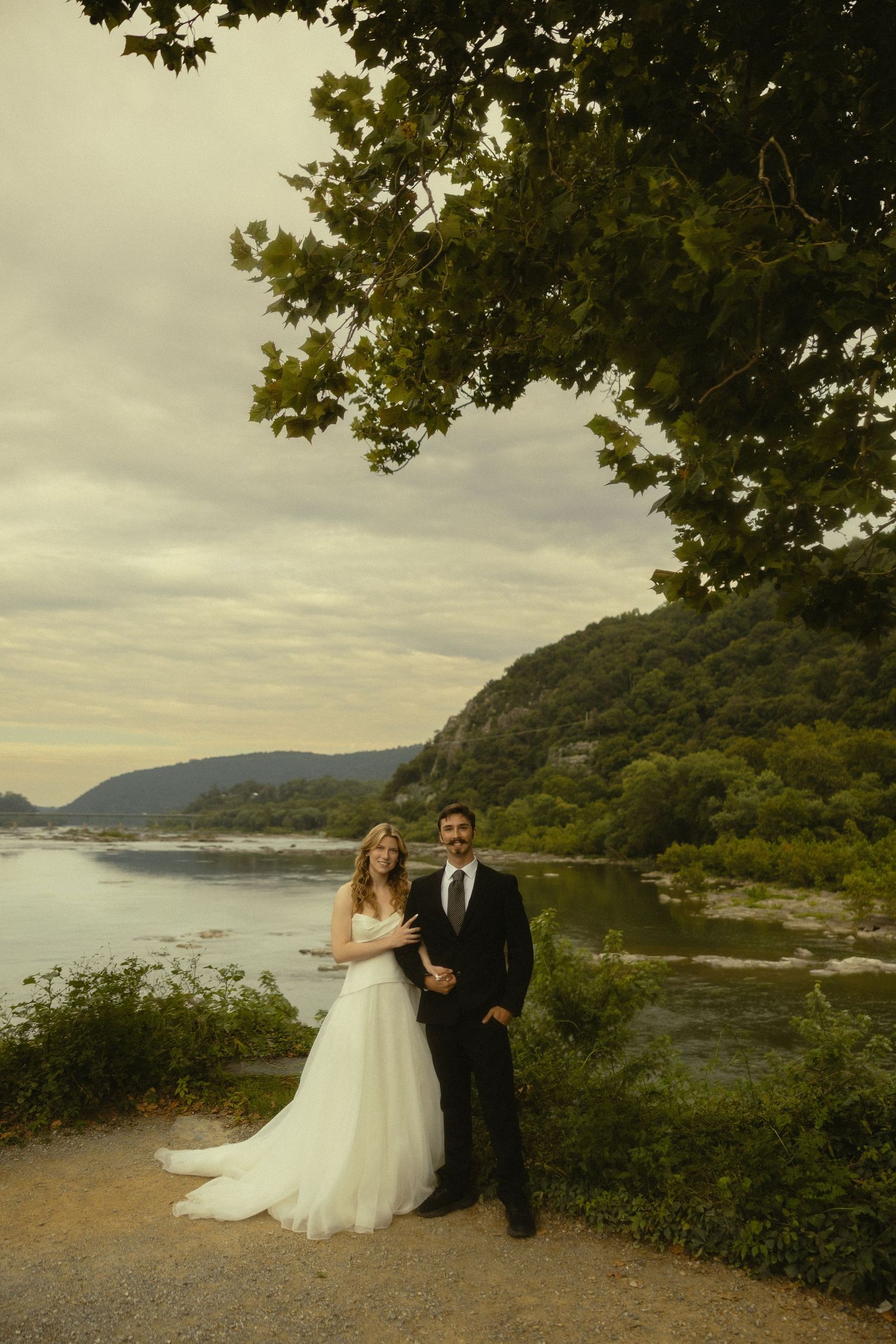 Bride and groom posing by a scenic river with lush green hills and trees at dusk on their wedding day.