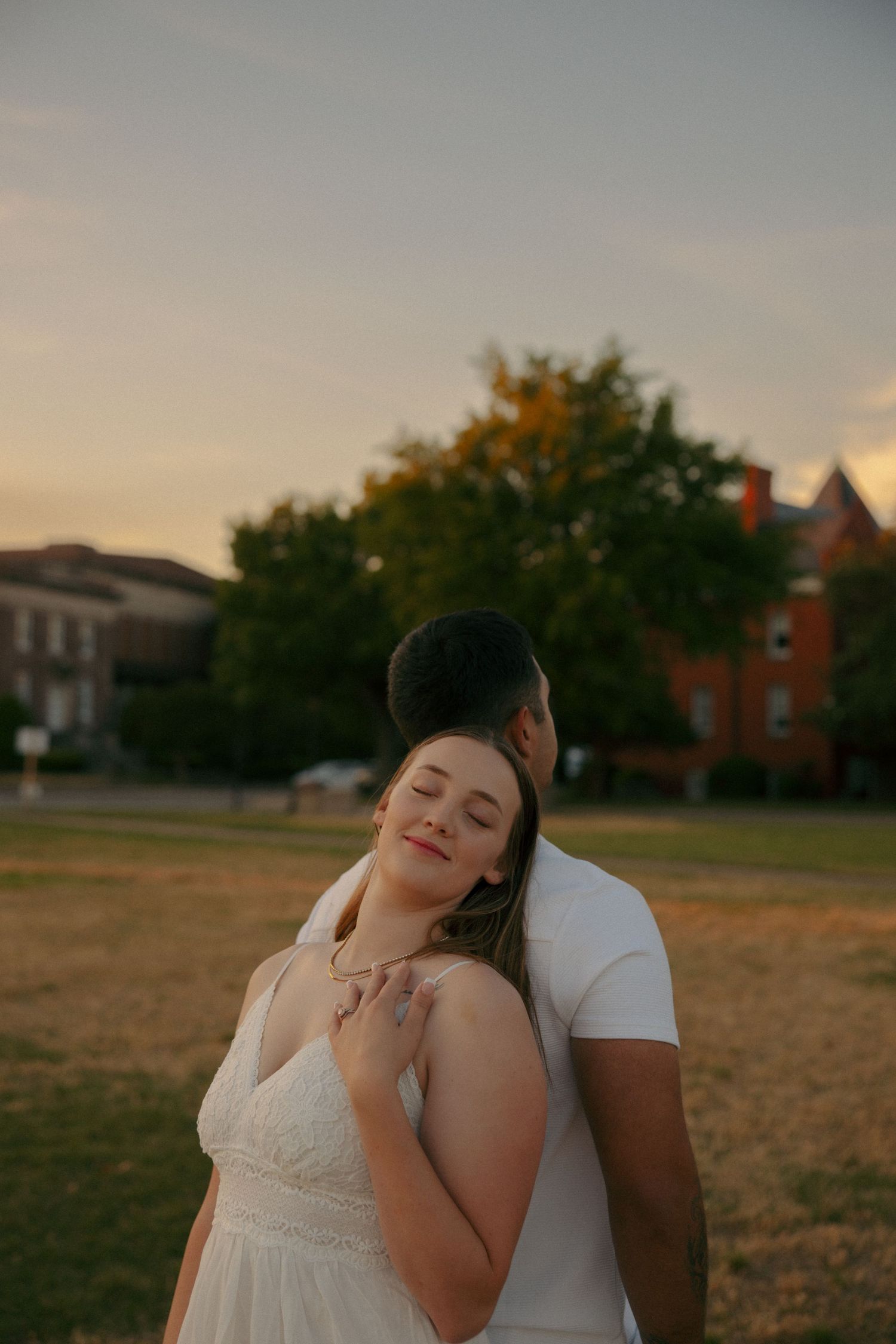 Couple embracing at sunset in a park, woman in white dress leaning back against man in white shirt, eyes closed.
