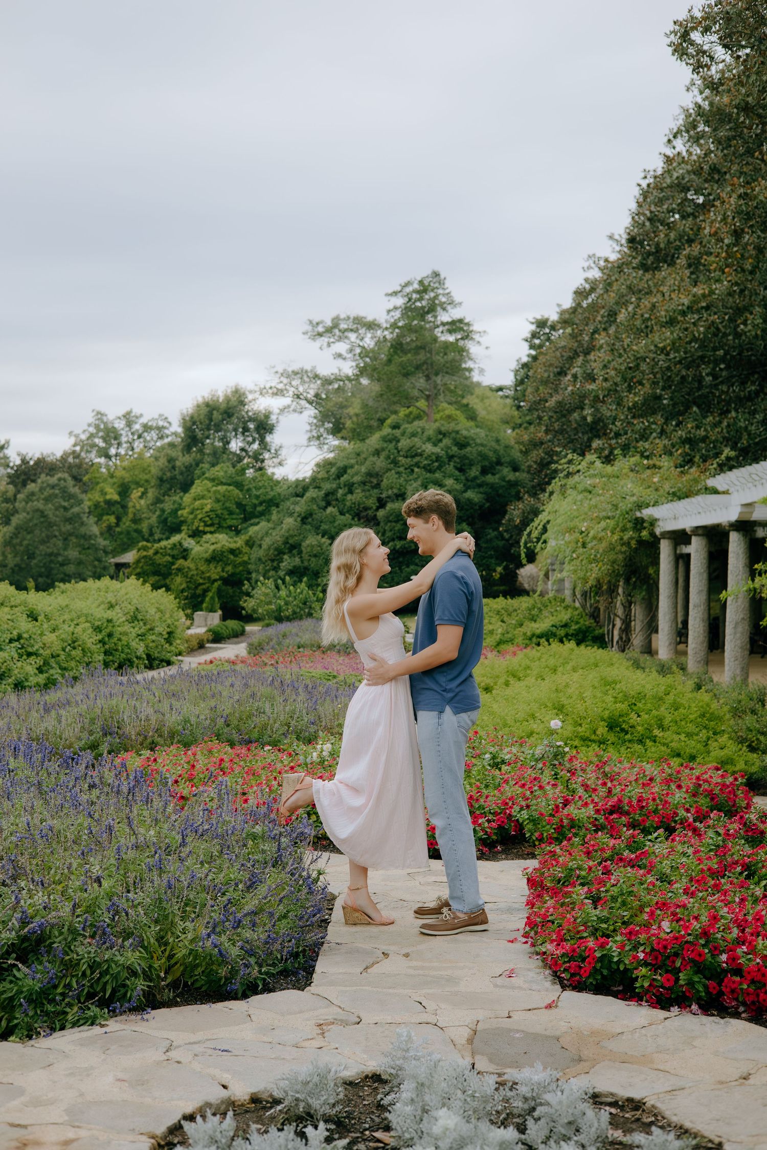 A couple embraces on a stone path surrounded by lavender and red flowers in a lush garden setting.