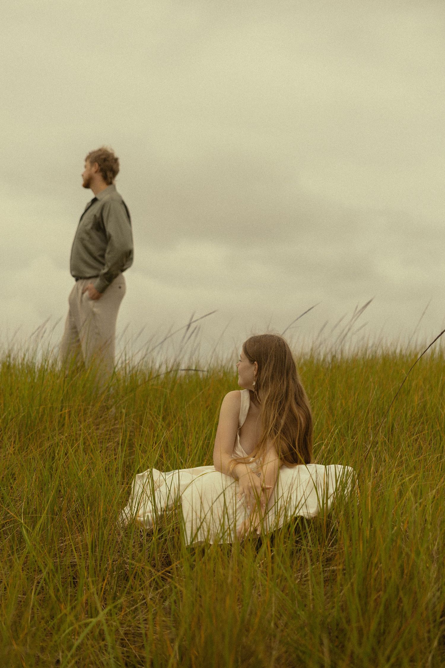 A woman in white dress sits in tall grass while a man stands behind her under overcast skies.
