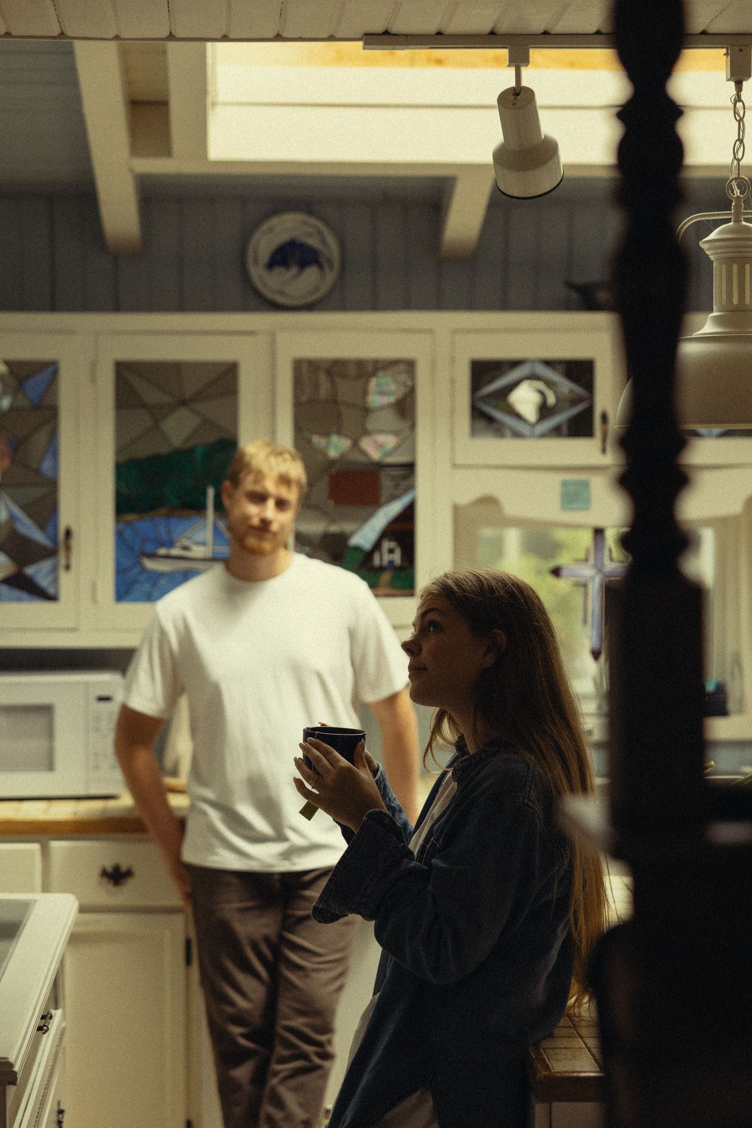 Couple relaxing in a cozy kitchen with stained glass cabinets, woman holding coffee cup, man in white t-shirt standing nearby.
