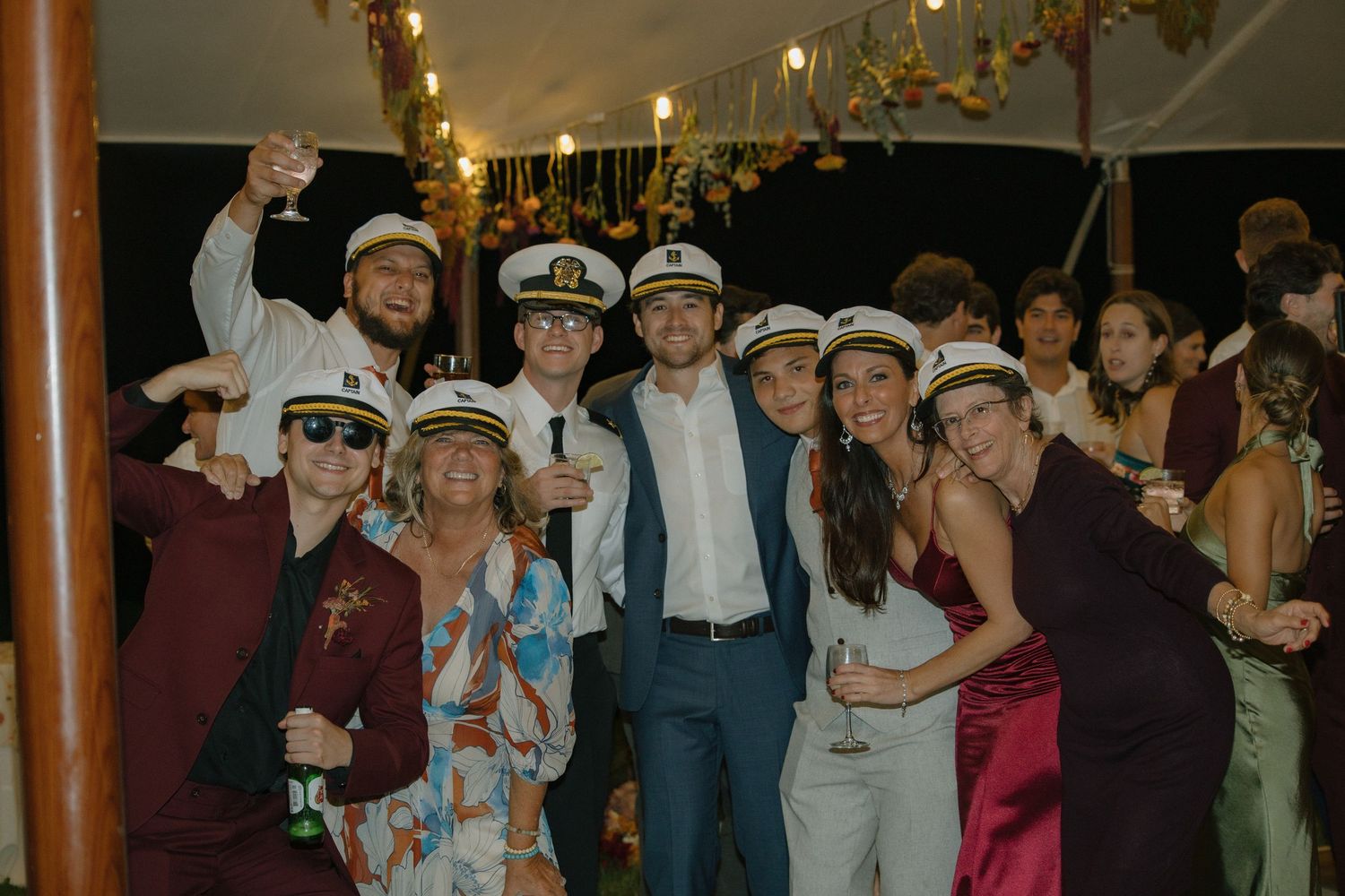 Wedding guests in sailor hats dance and celebrate at a reception party with chandelier lighting overhead.