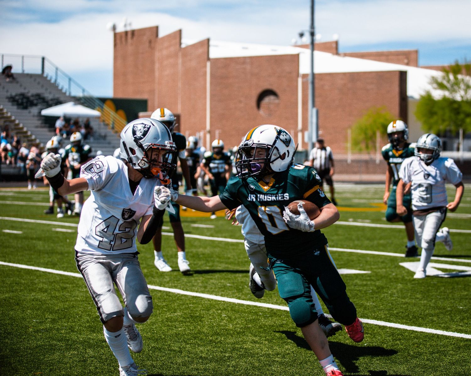 Youth football players compete on a sunny field, with a ball carrier in teal uniform evading a defender in white.