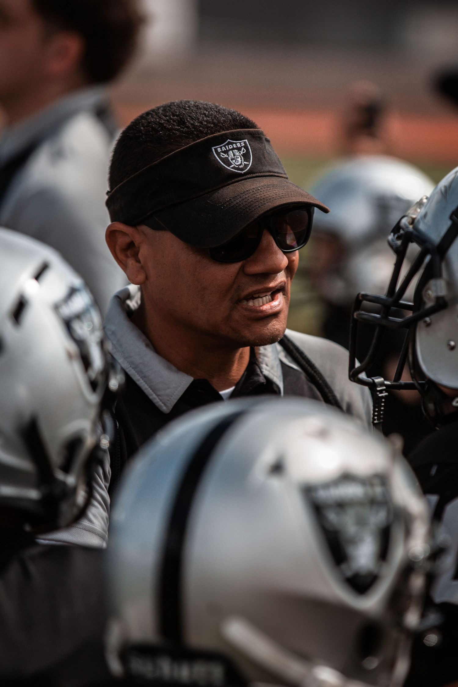 Raiders coach wearing black visor and sunglasses talks to players in silver helmets on the football field.