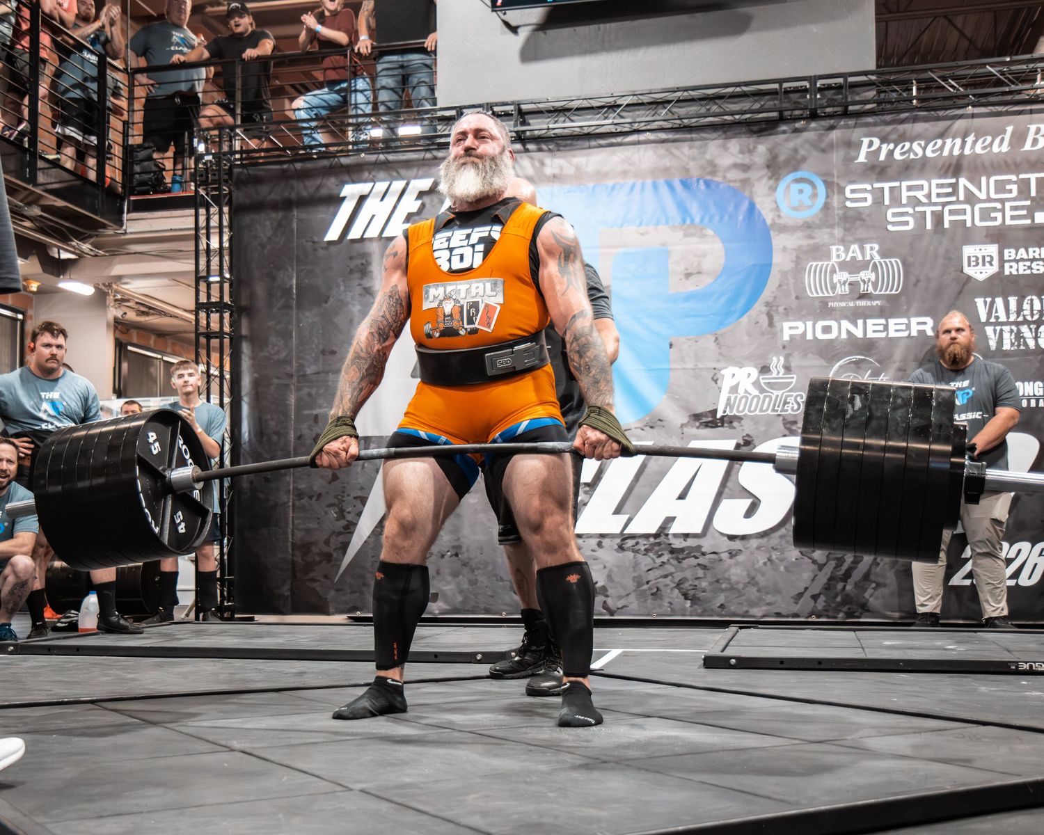 Athletes competing in powerlifting event with orange shirts and professional staging setup.
