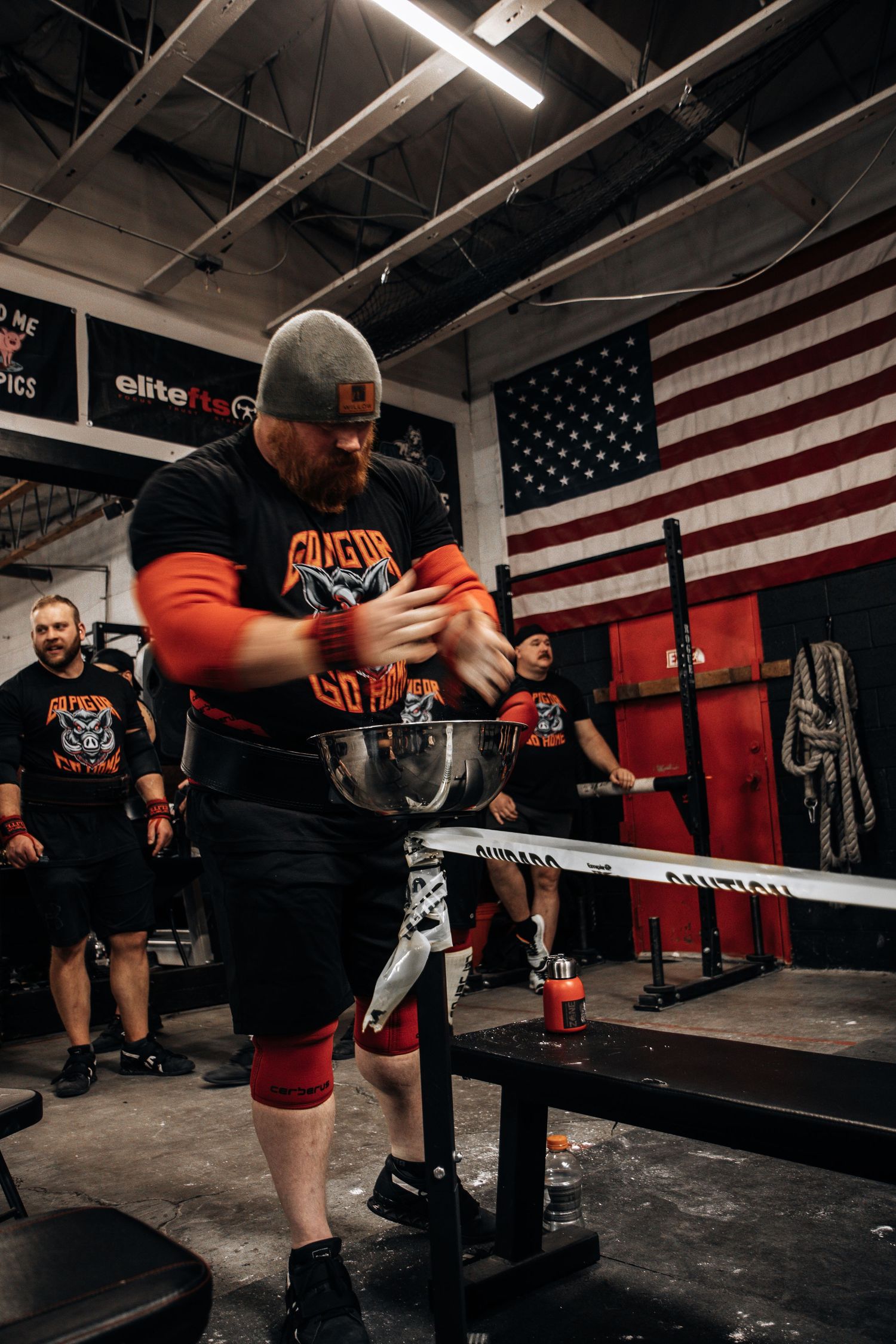 Athletes perform intense weightlifting exercises in a gym with an American flag backdrop.