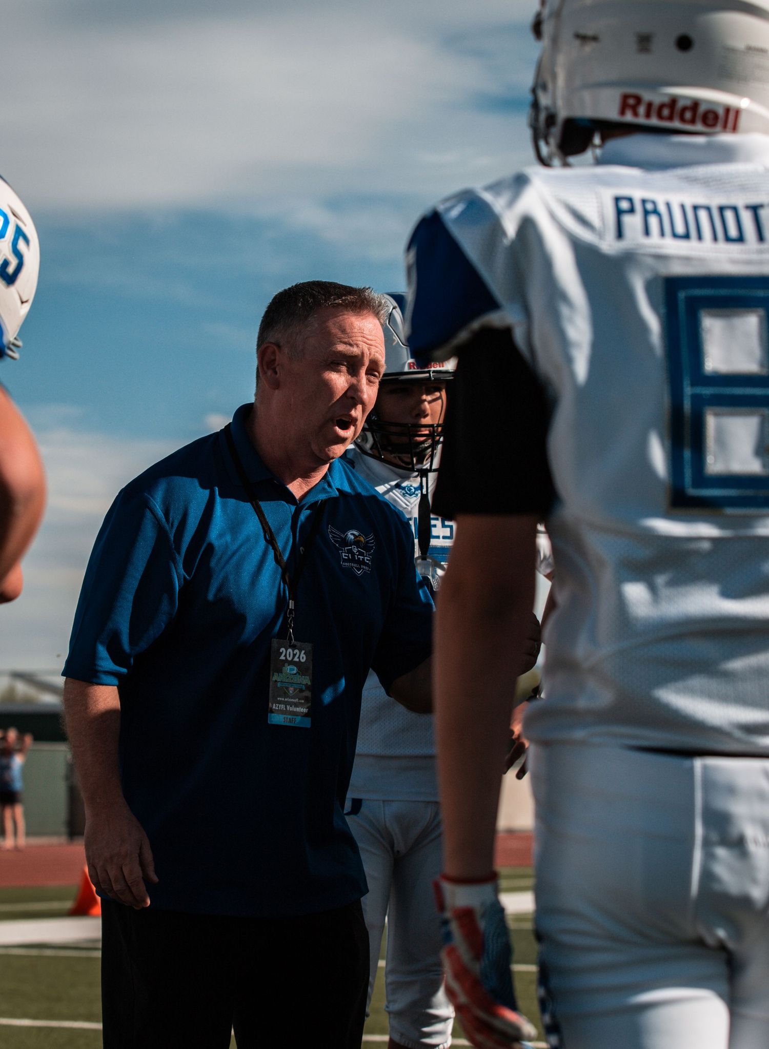 A football coach in a blue polo talks to player wearing Riddell helmet and Prunot jersey on the sideline.