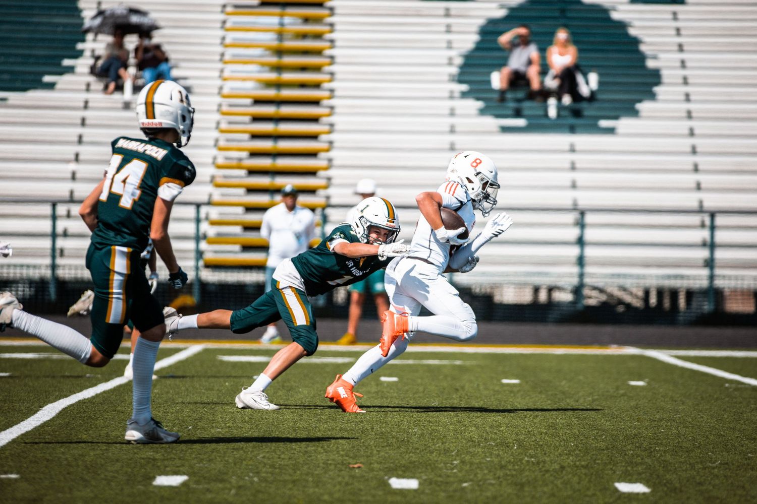 Football players compete on a green field, with one player in white evading a tackle during a sunny game.