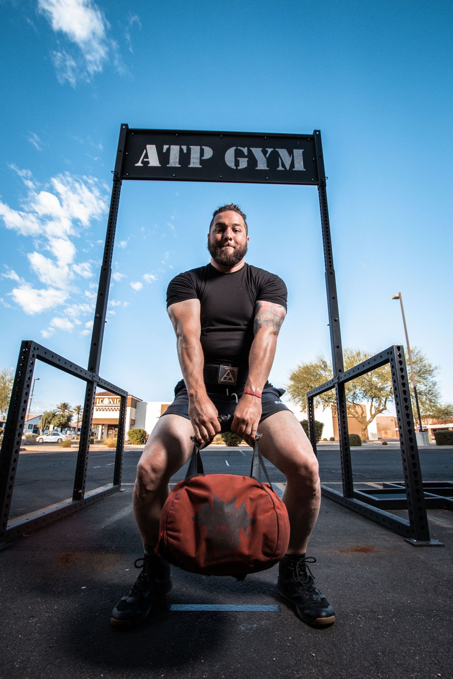 Muscular man lifting a red kettlebell outdoors at ATP Gym under a blue sky.