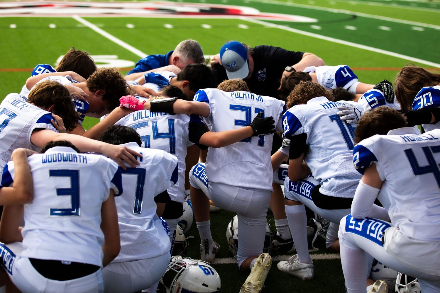 Youth football team huddles together in prayer on field, wearing blue and white uniforms after a game.