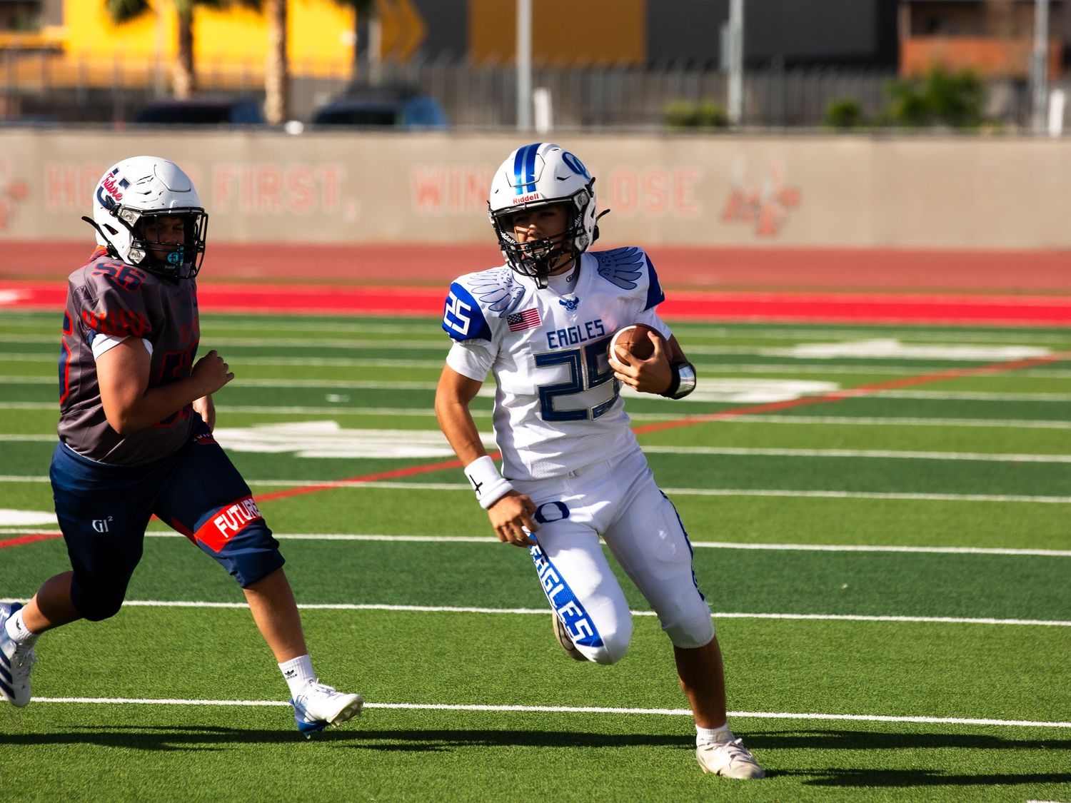 Eagles football player #25 in blue uniform runs with the ball during a high school football game on red turf.