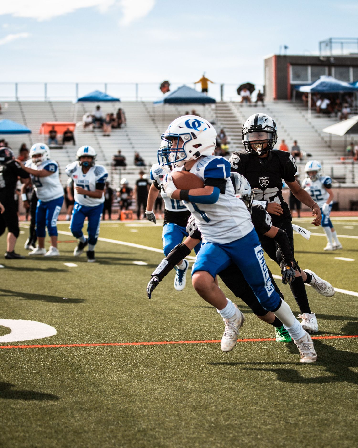 Youth football player in blue uniform runs with ball during a game at a stadium with fans in the stands.
