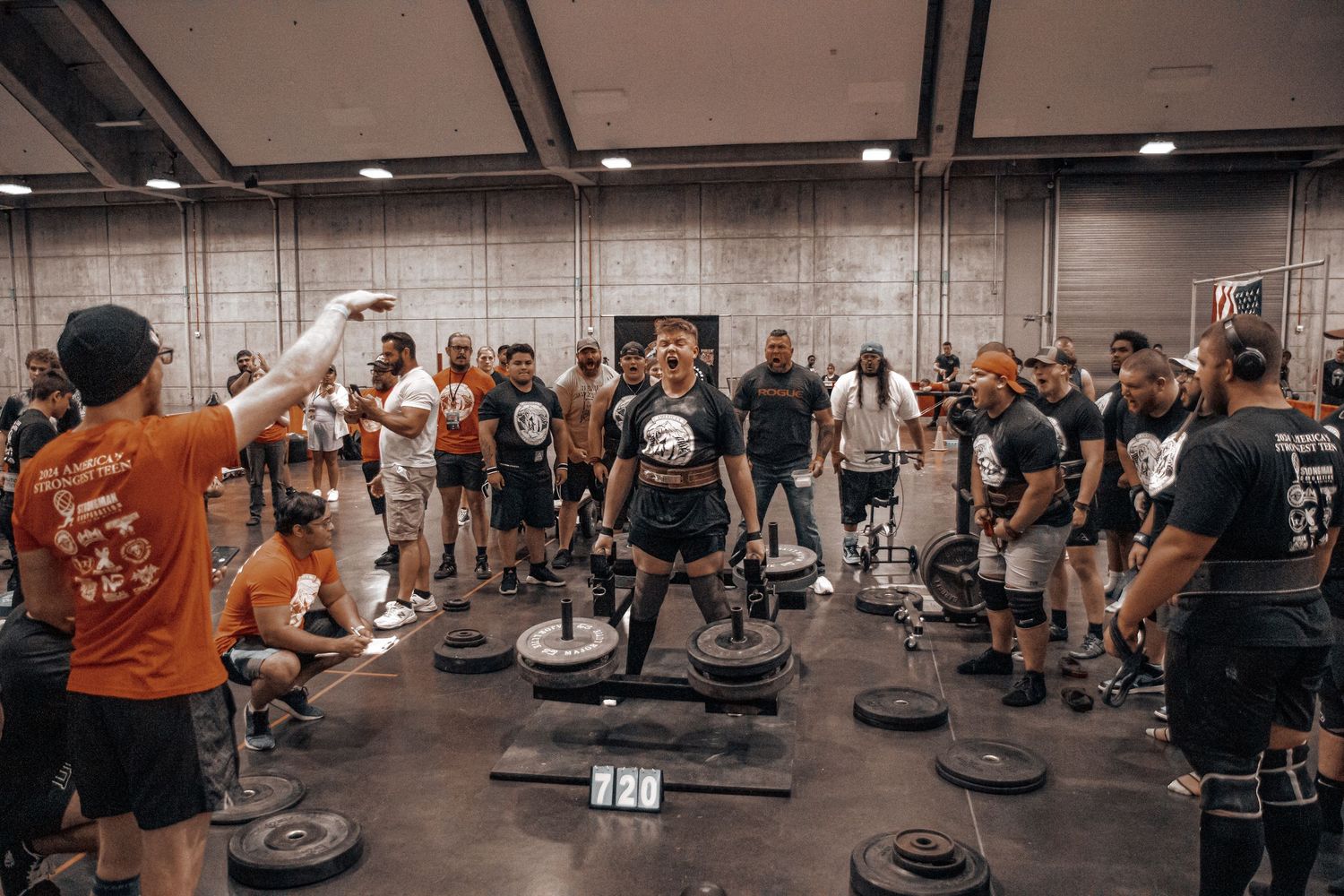 Group fitness class participants exercise with weights in a spacious industrial gym setting.