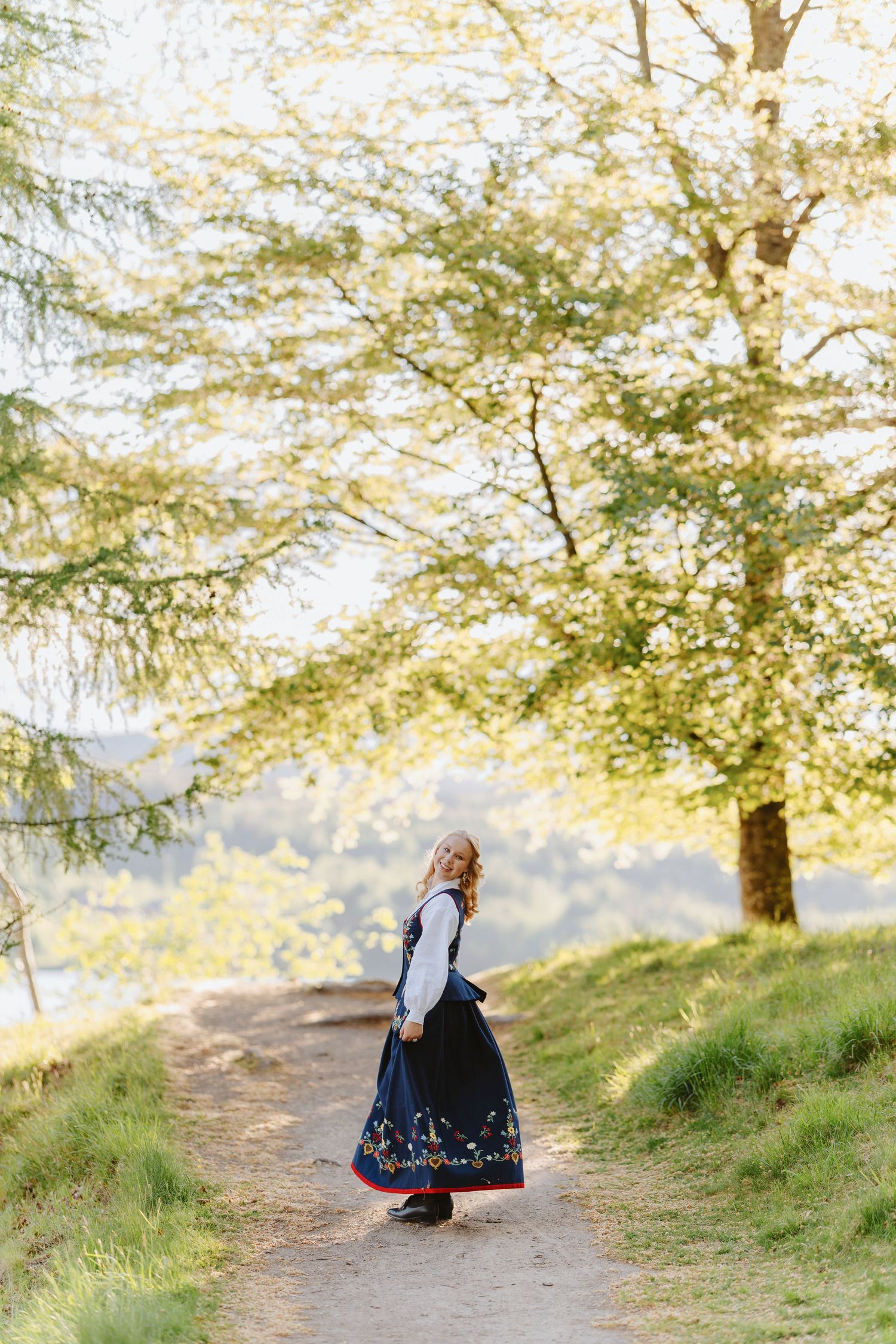 Woman in traditional dirndl dress standing on a scenic forest path surrounded by lush green trees in soft sunlight.