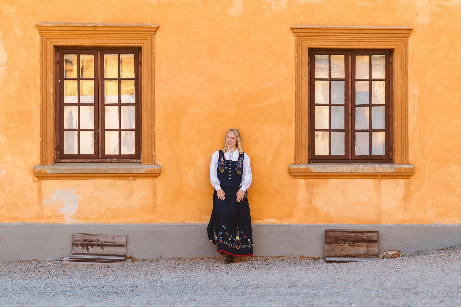 Woman in traditional Scandinavian dress stands against vibrant orange building with wooden-framed windows.