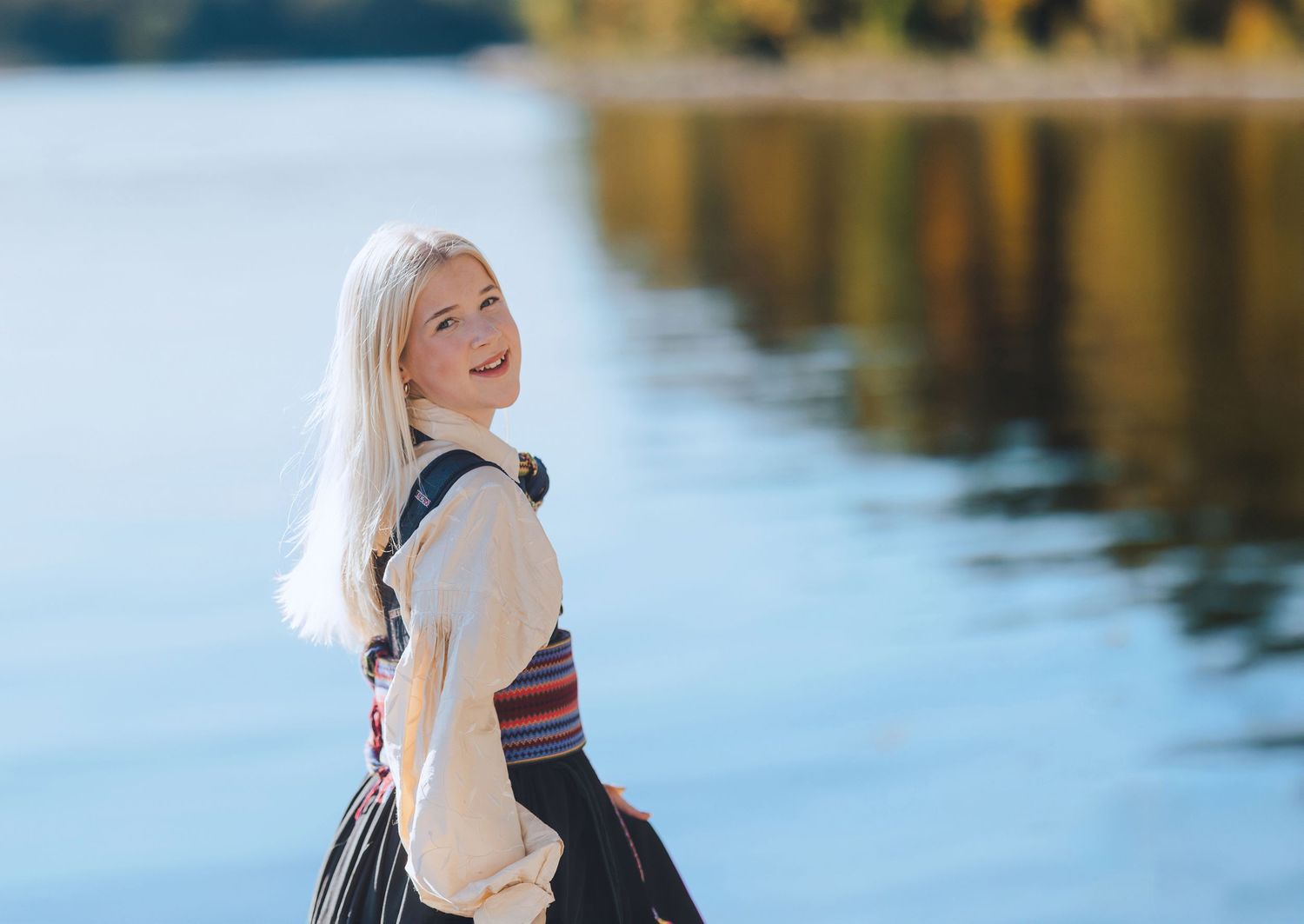 Woman in white sweater smiling and posing outdoors by a reflective lake during autumn season.
