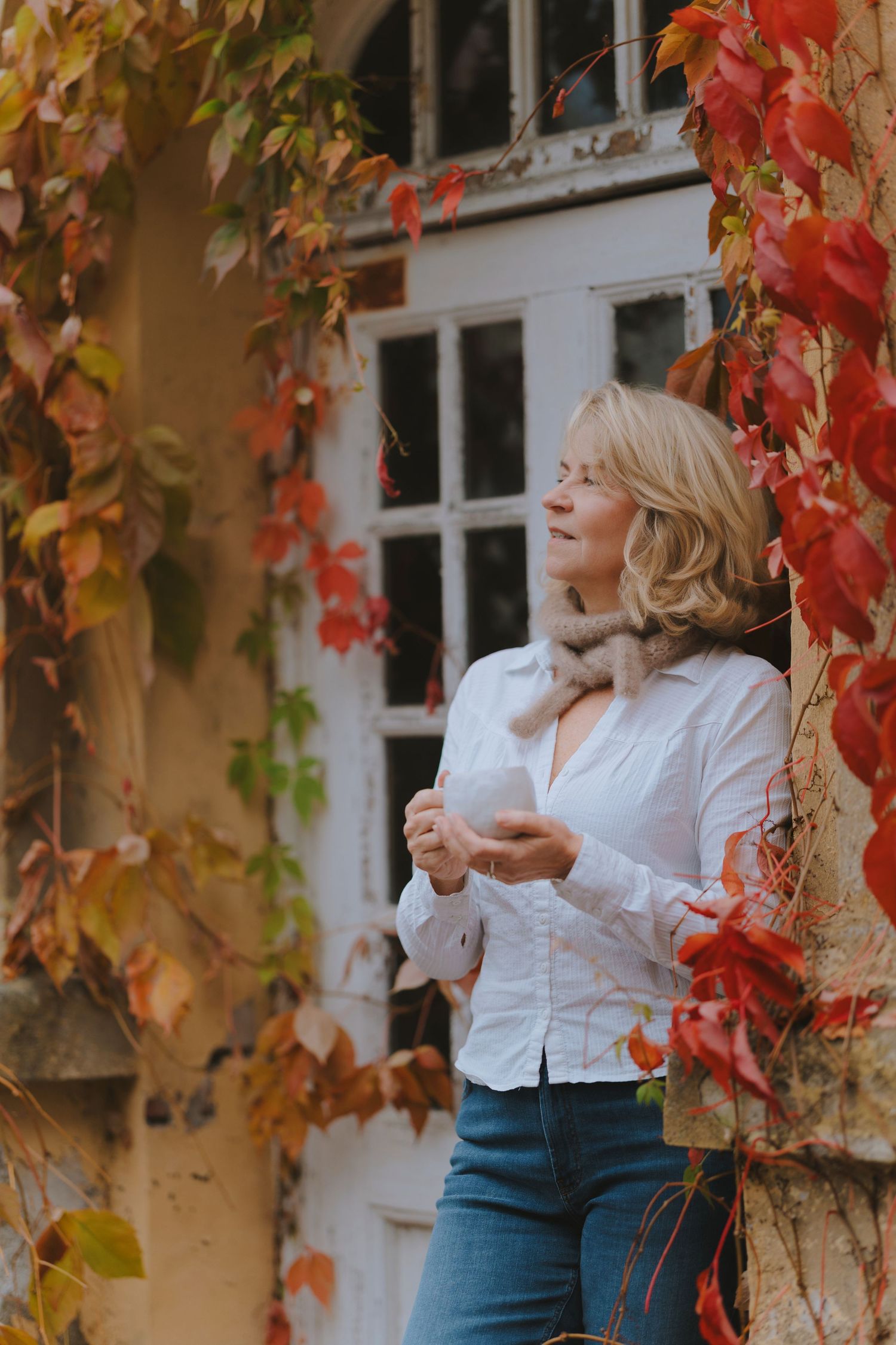 Woman in white button-down shirt stands against autumn wall with vibrant red ivy leaves in natural lighting.